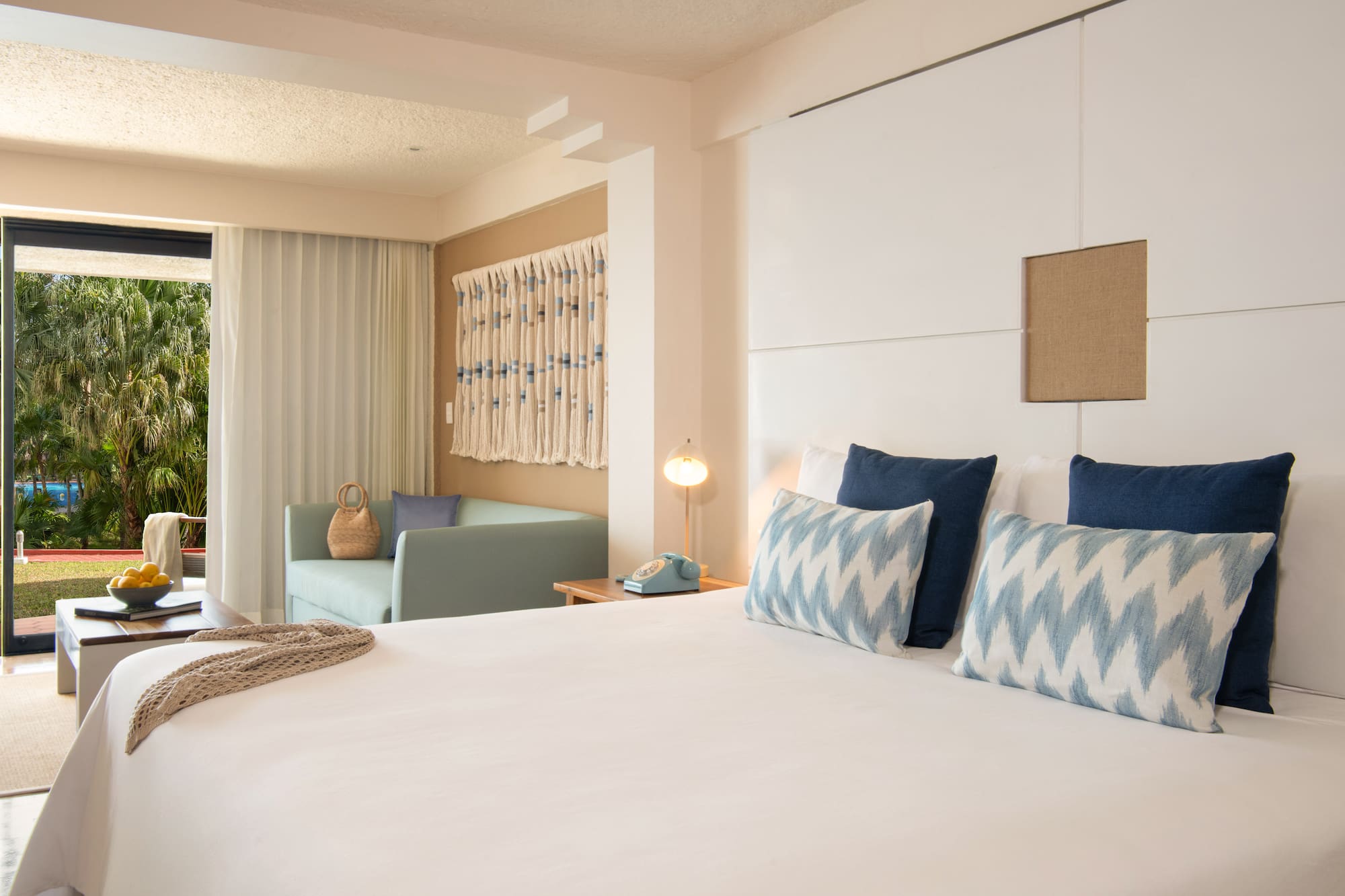 Hotel room with bed, blue pillows, sofa, and tropical palm garden view.