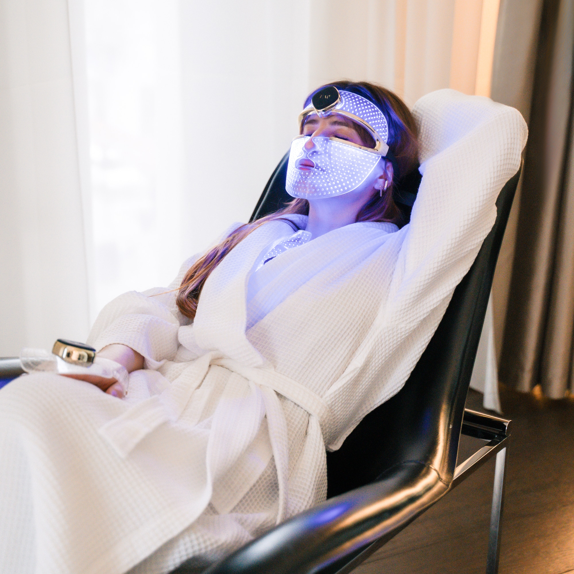 Relaxing woman in bathrobe with blue LED light therapy mask for facial skincare.