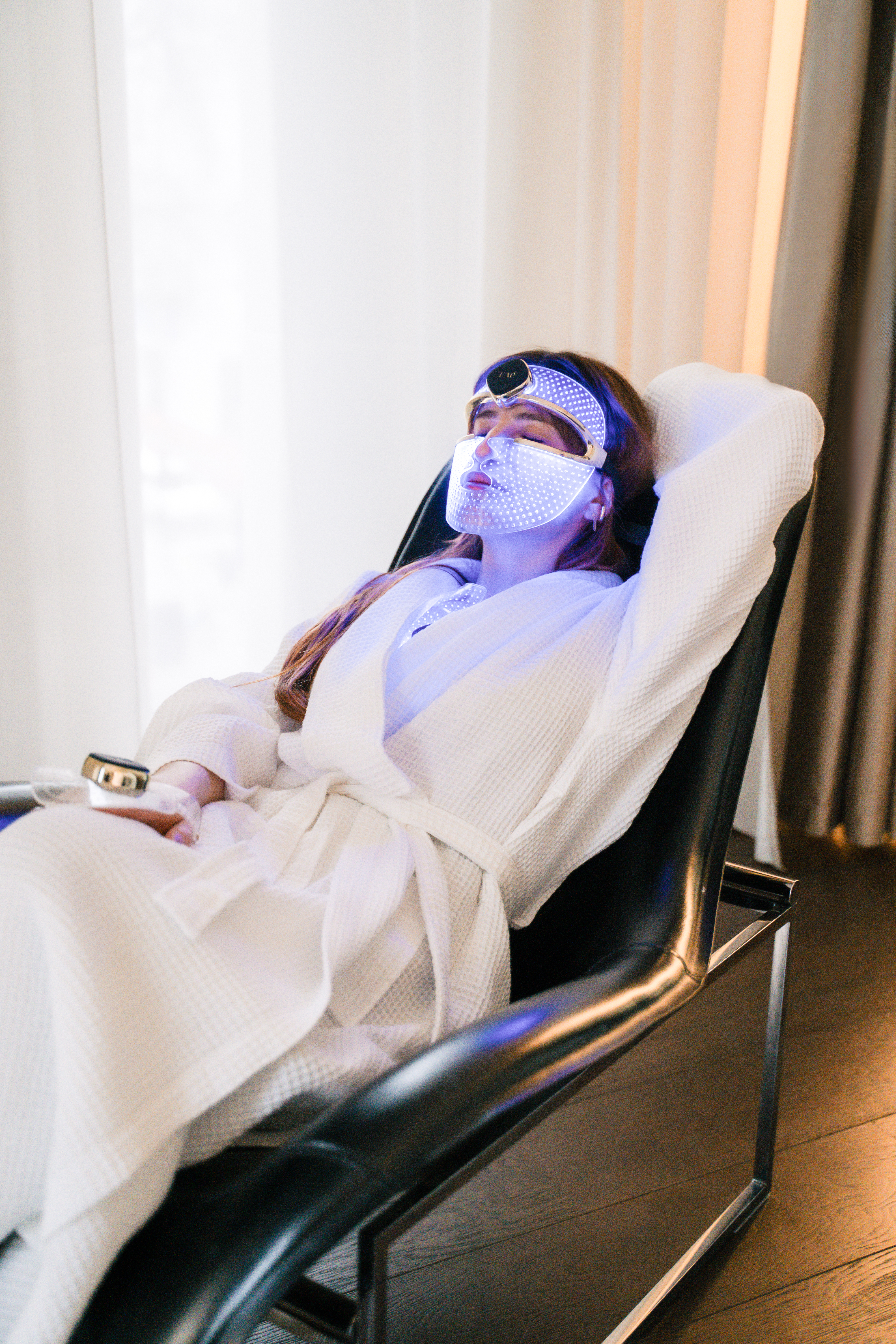 Relaxing woman in bathrobe with blue LED light therapy mask for facial skincare.