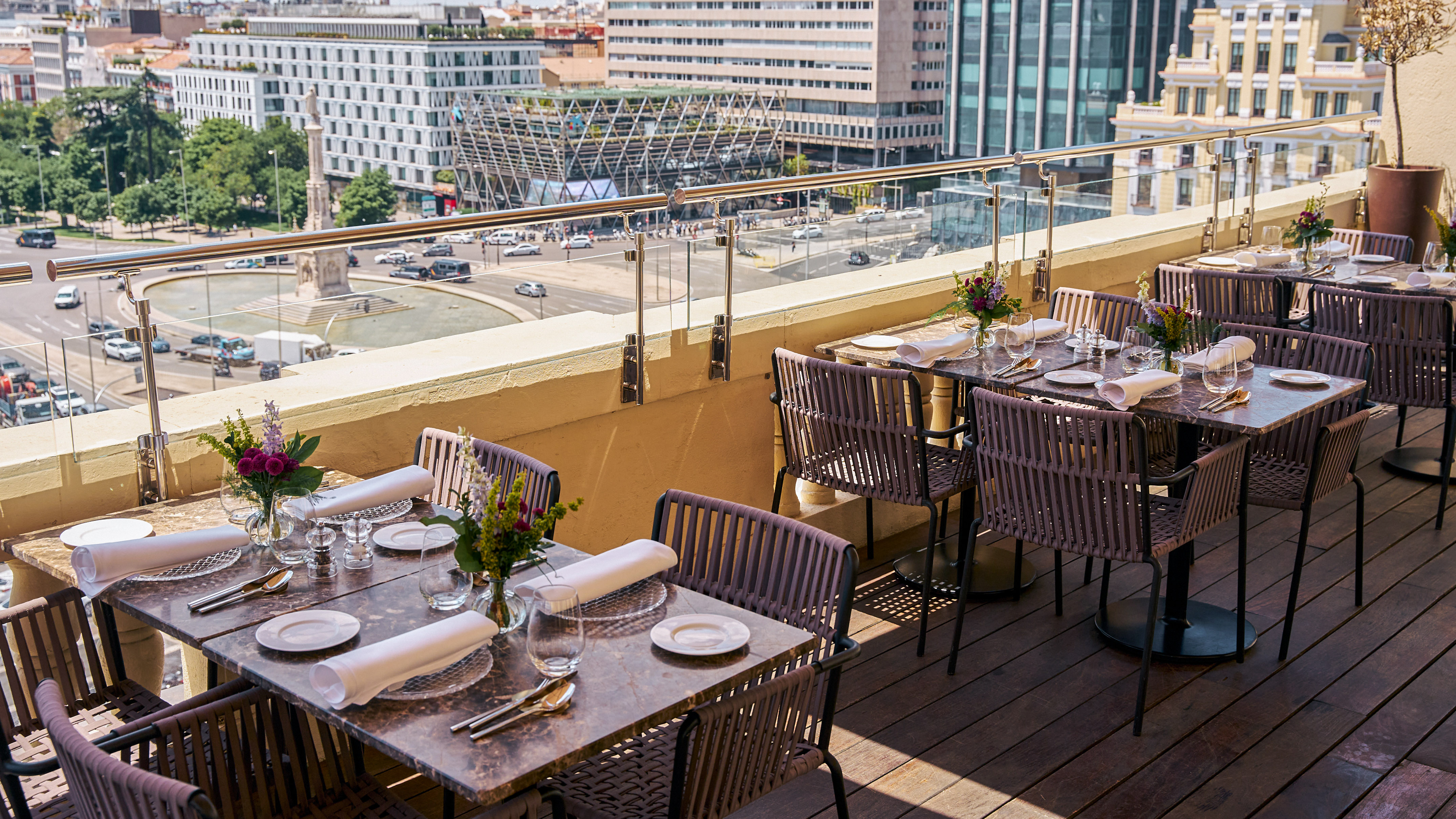 Outdoor rooftop dining terrace with set tables, woven chairs, and cityscape view.