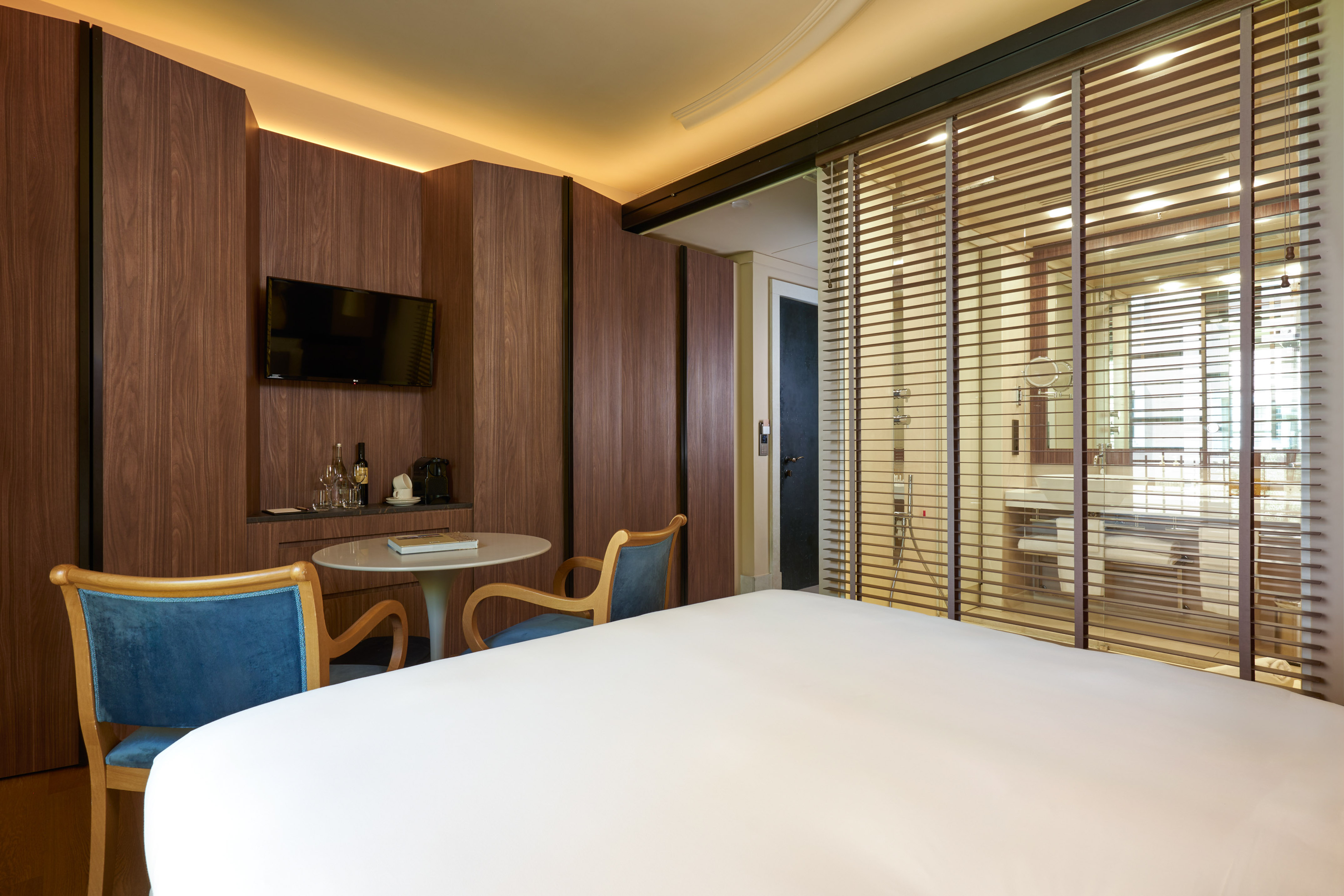 Modern hotel room, wood paneling, bed, TV, and bathroom behind slatted blinds.