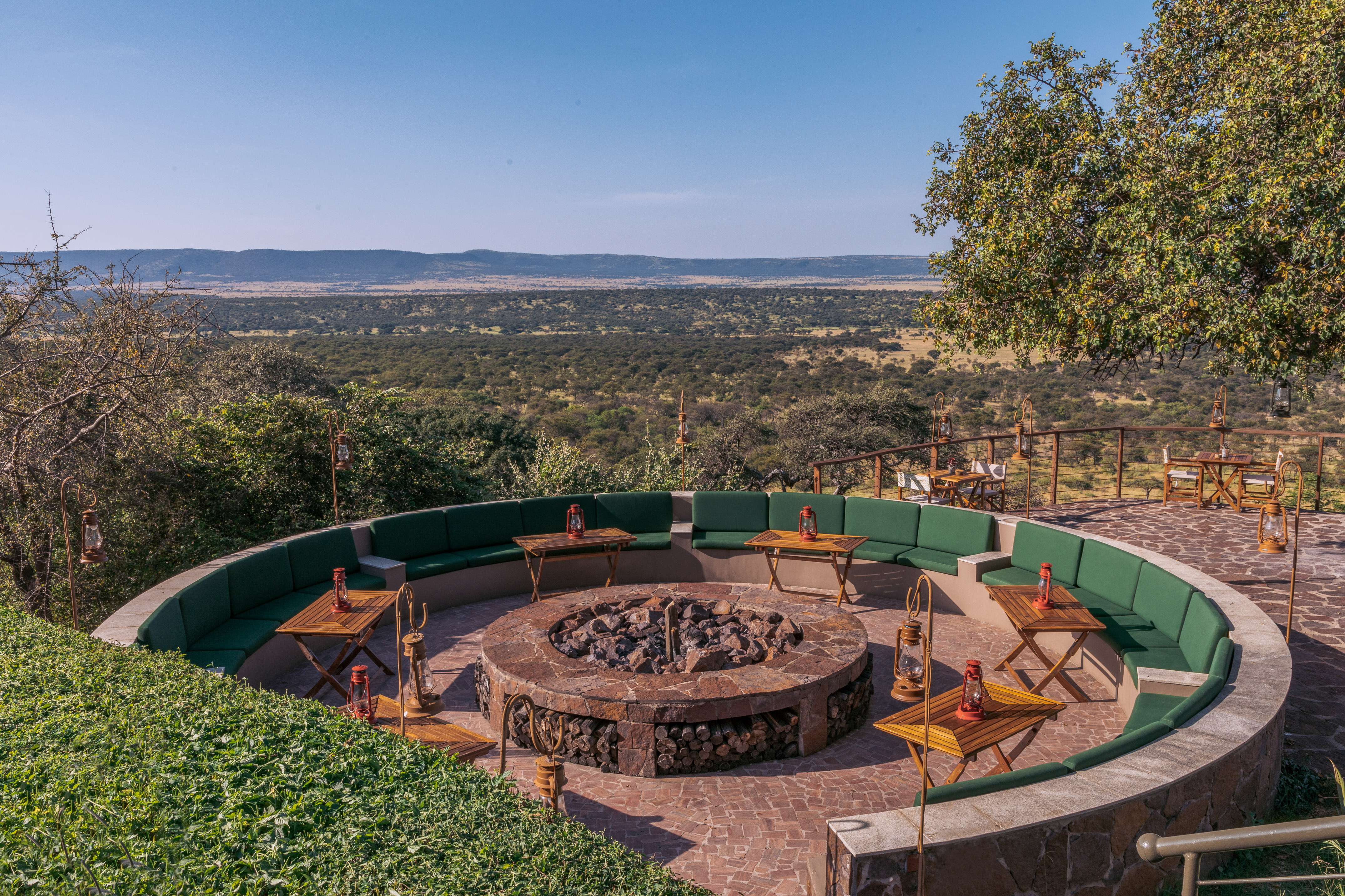 Safari lodge fire pit lounge, green seating, savanna landscape view.