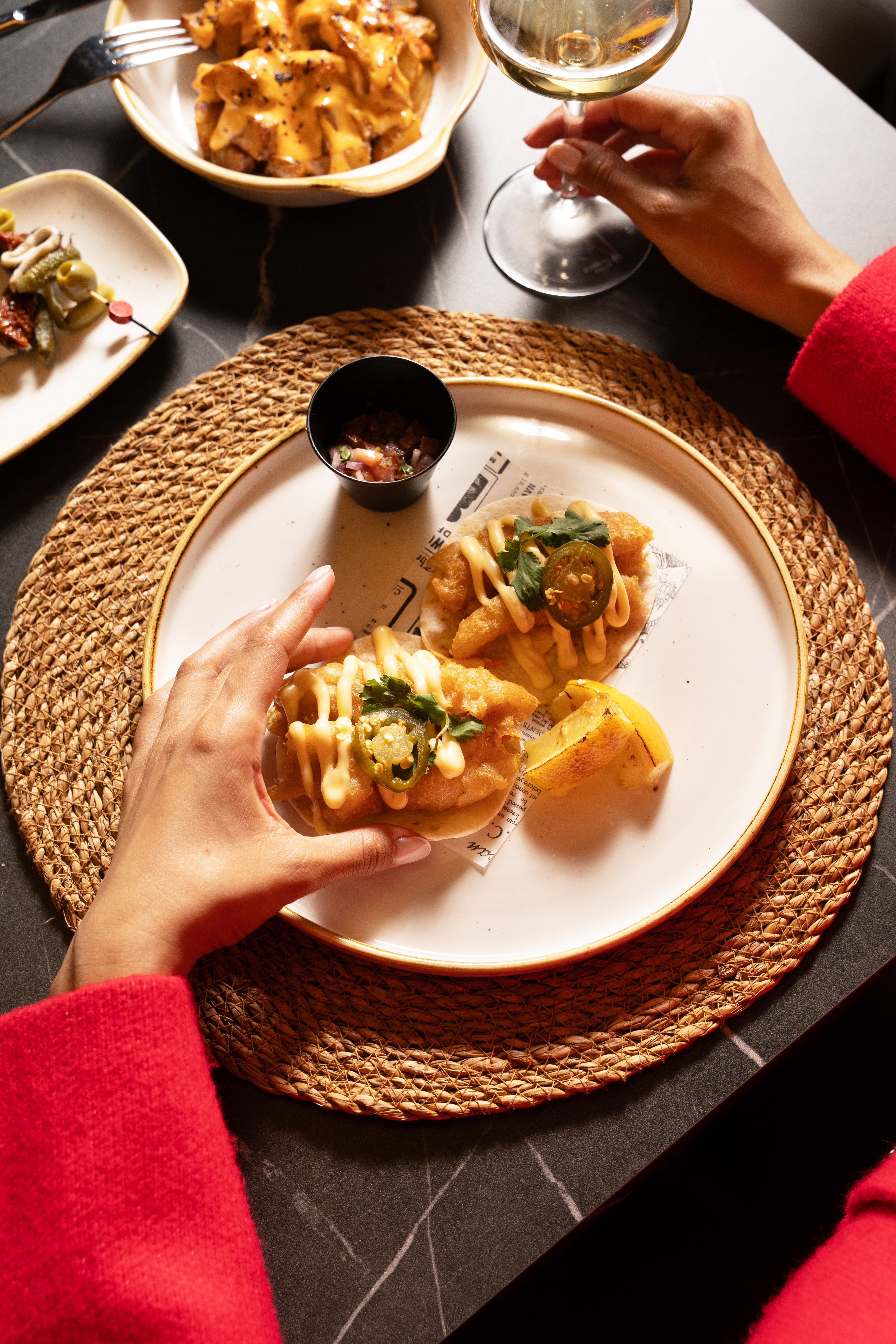 Person grabbing fish tacos with jalapeños, salsa, wine, and patatas bravas.