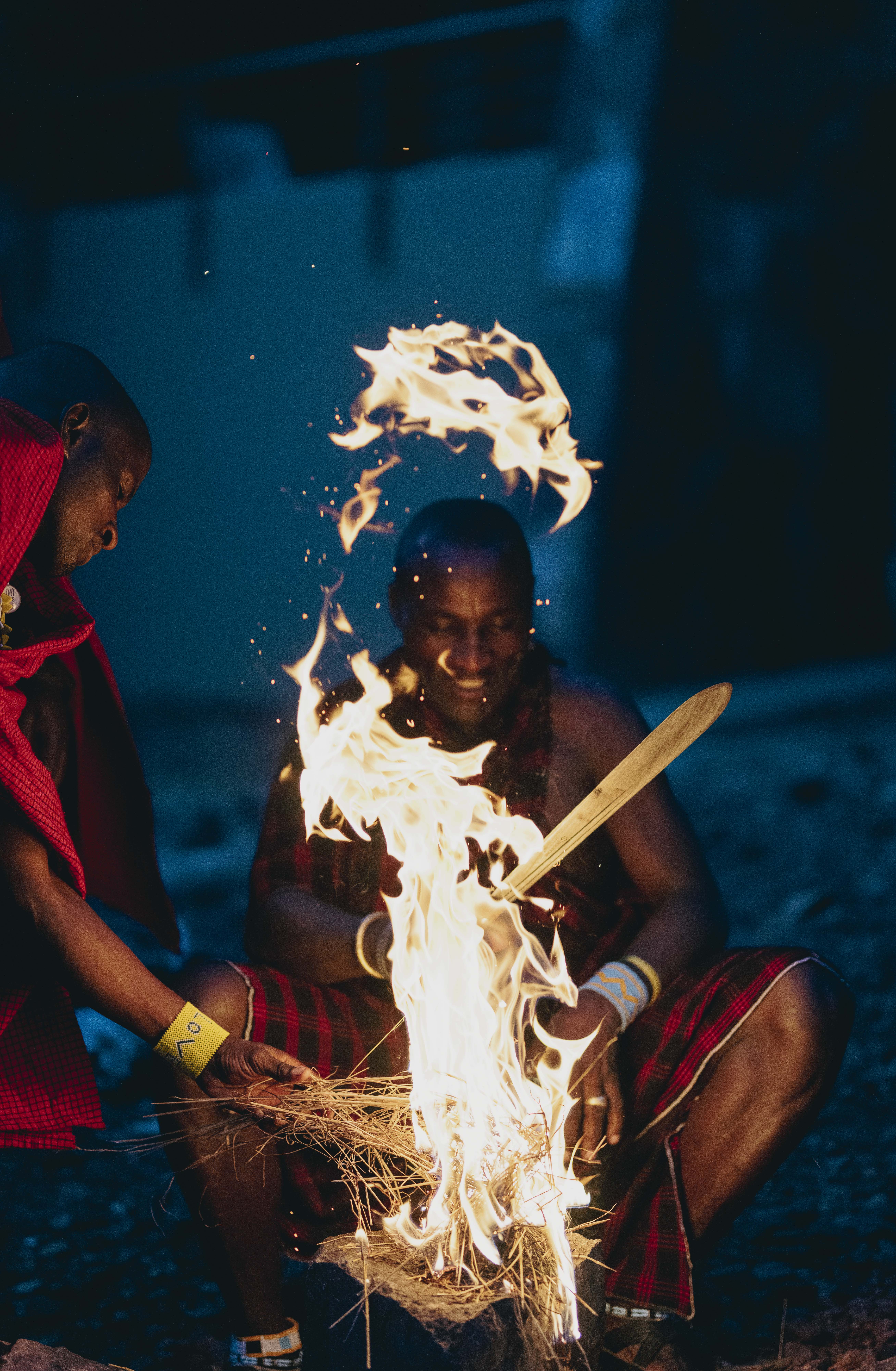 Maasai warriors starting a traditional fire at night, one adding kindling.