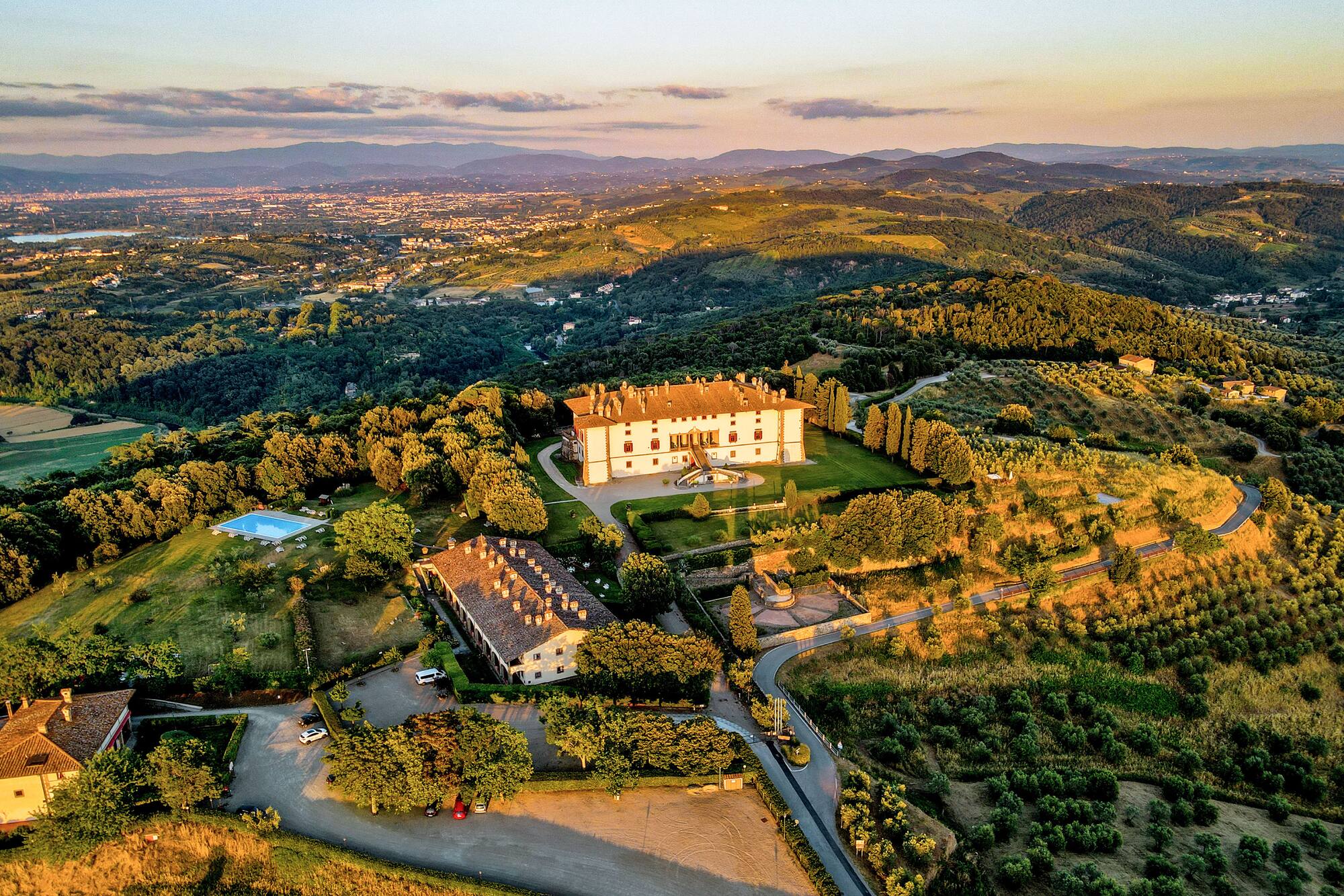Aerial view: Tuscan villa estate, pool, vineyards, and hills at golden hour.