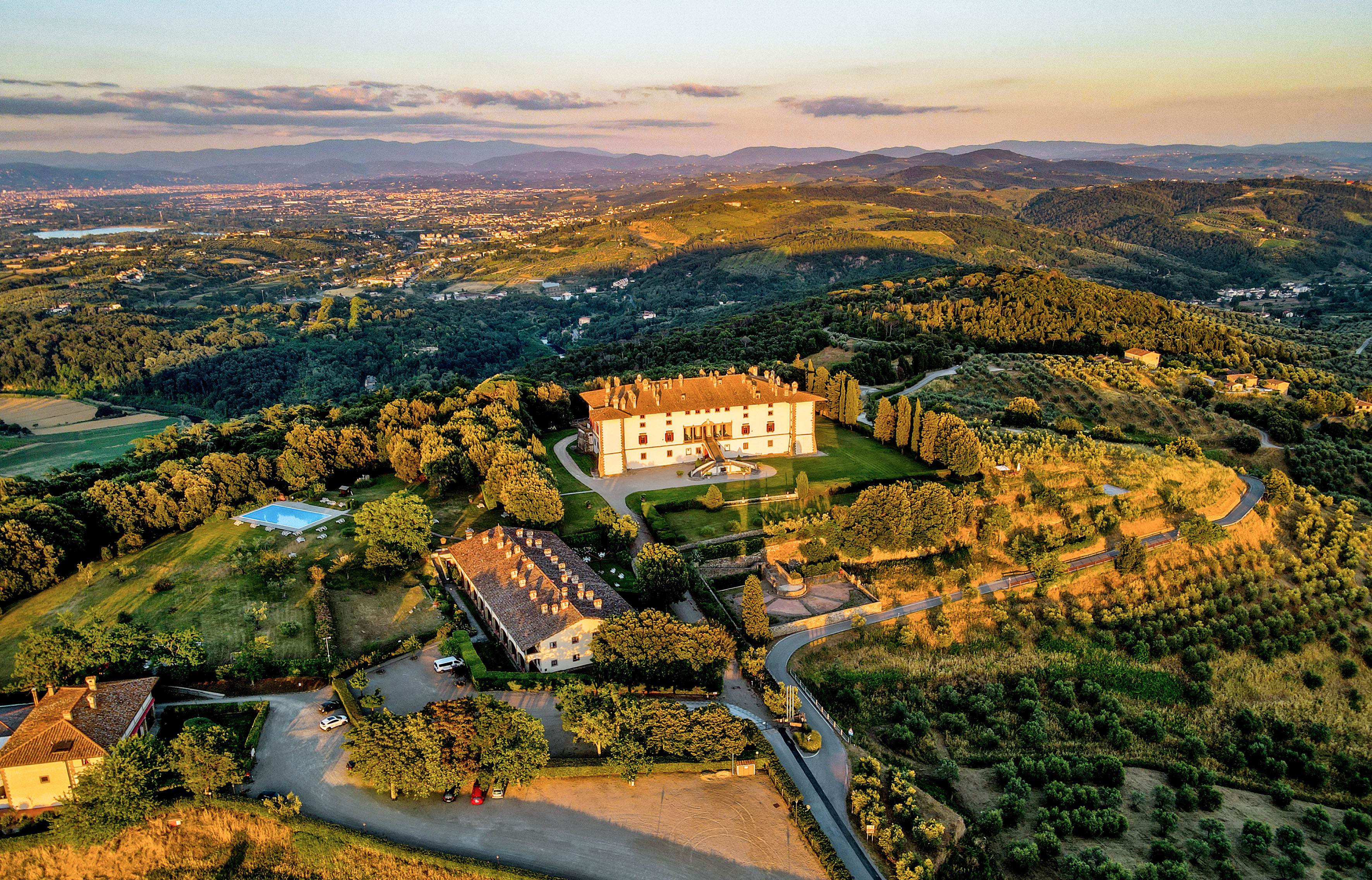Aerial view: Tuscan villa estate, pool, vineyards, and hills at golden hour.