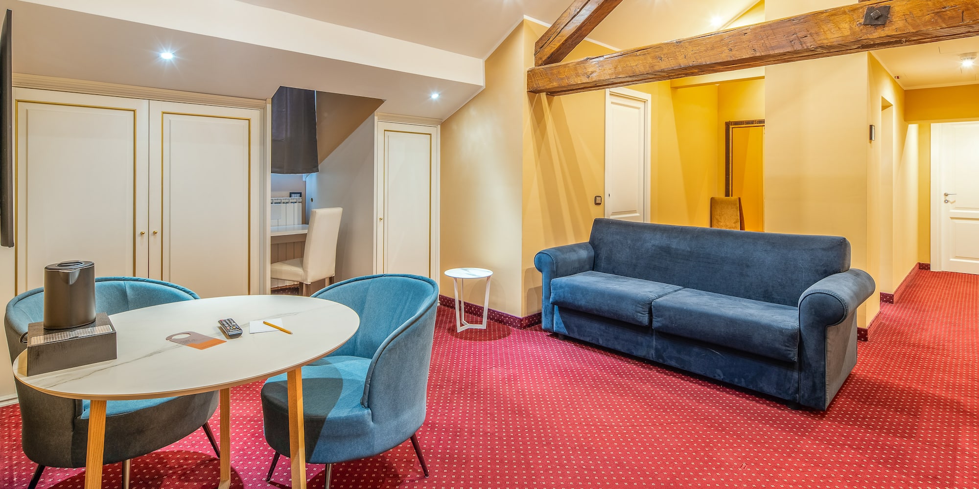 Hotel suite living room with red patterned carpet, blue sofa, and wooden beams.