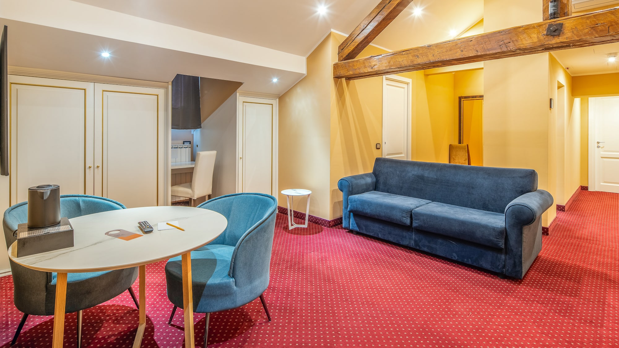 Hotel suite living room with red patterned carpet, blue sofa, and wooden beams.