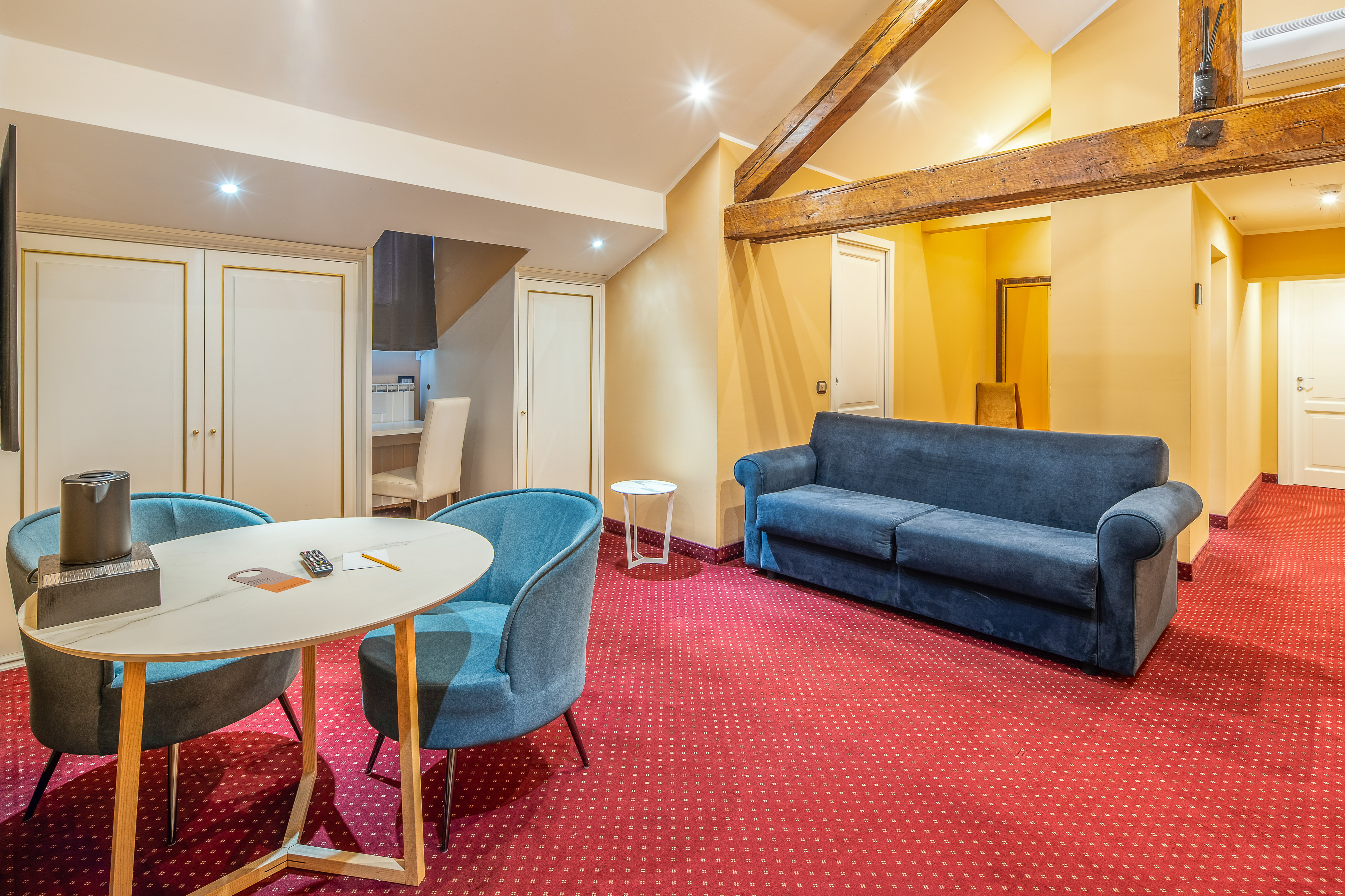 Hotel suite living room with red patterned carpet, blue sofa, and wooden beams.