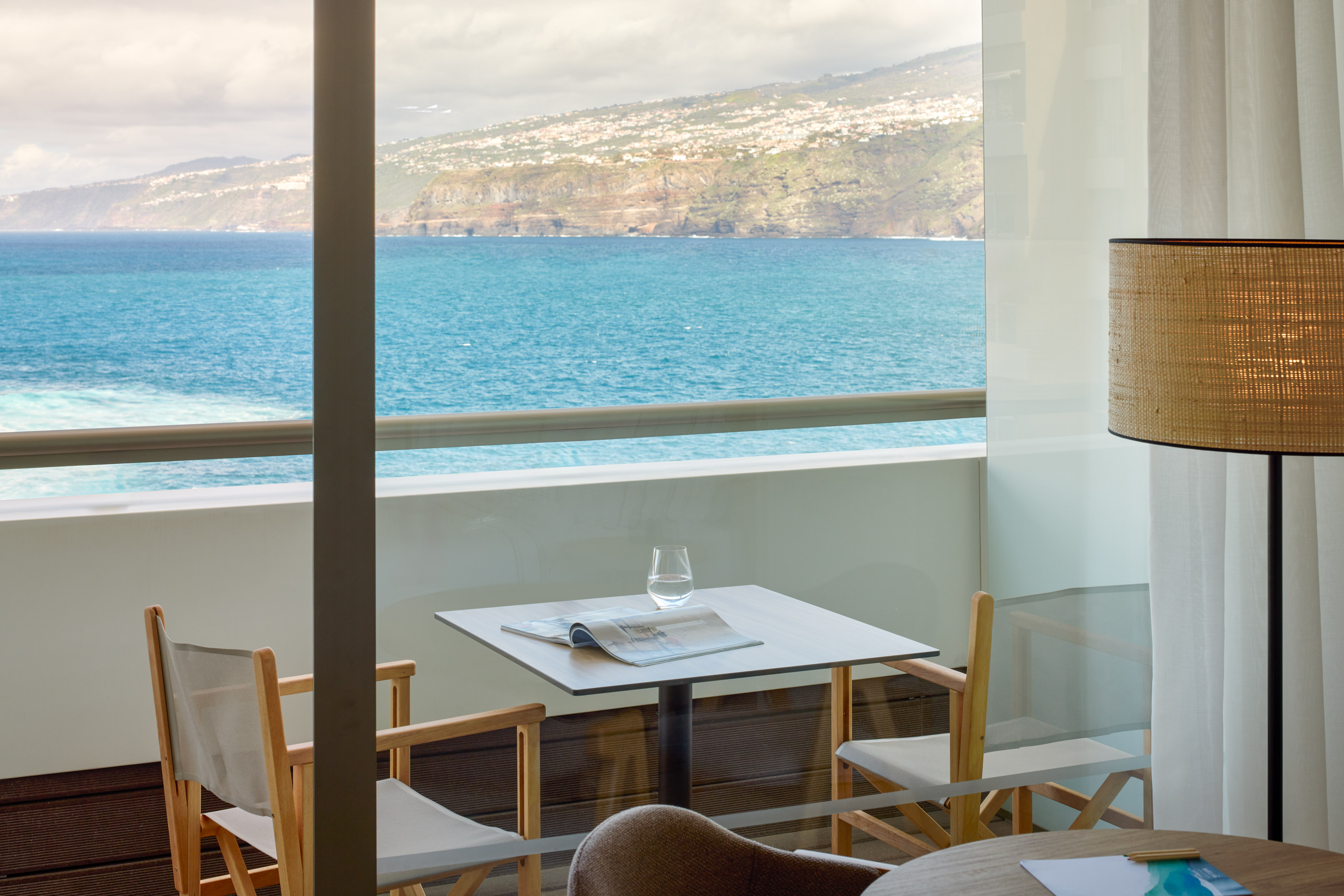 Coastal hotel balcony with ocean view, outdoor table, chairs, magazine, glass.