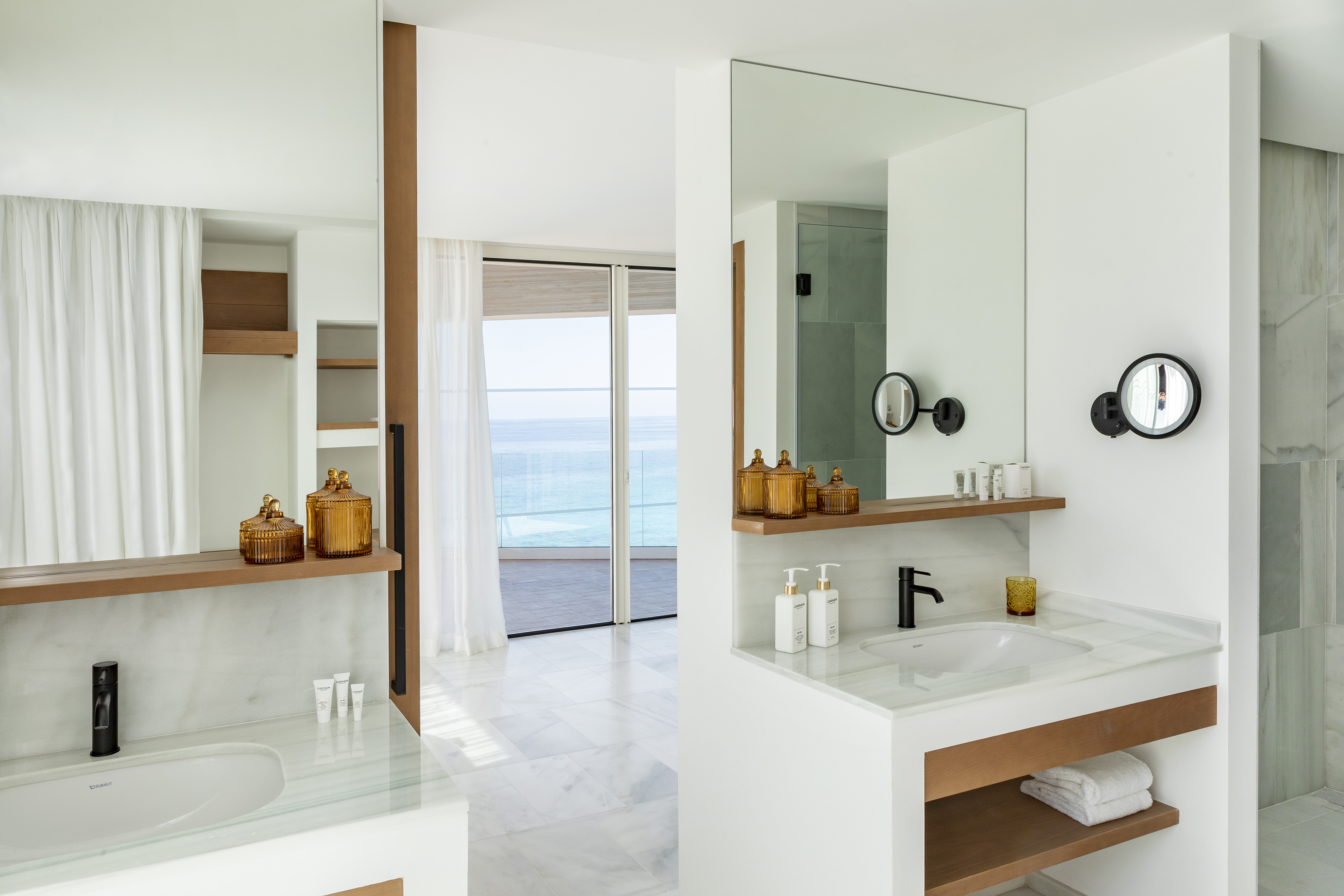 Modern hotel bathroom with ocean view, marble vanity, wooden shelf, and mirror.
