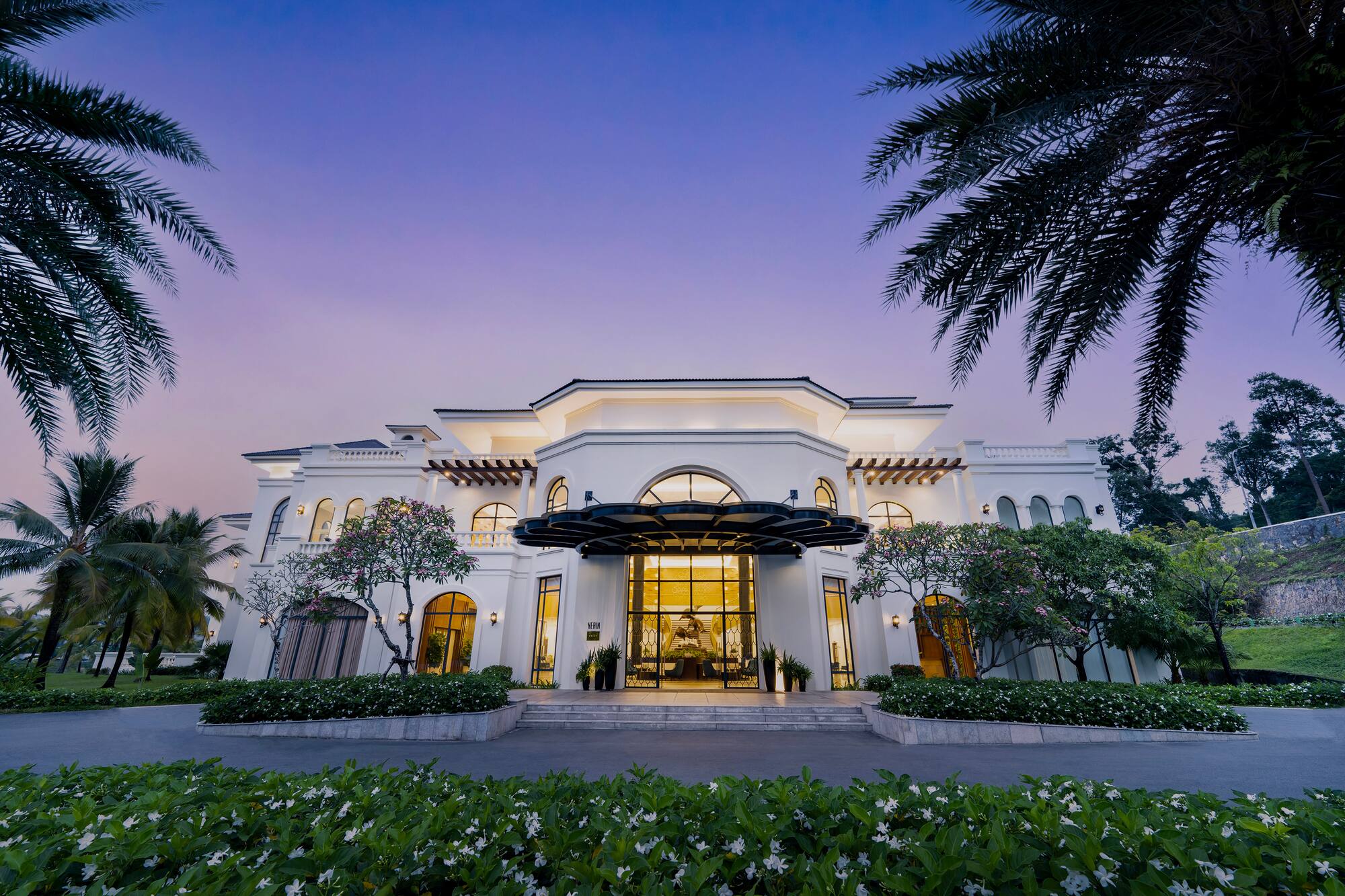 Grand white luxury hotel entrance with palm trees at purple dusk.