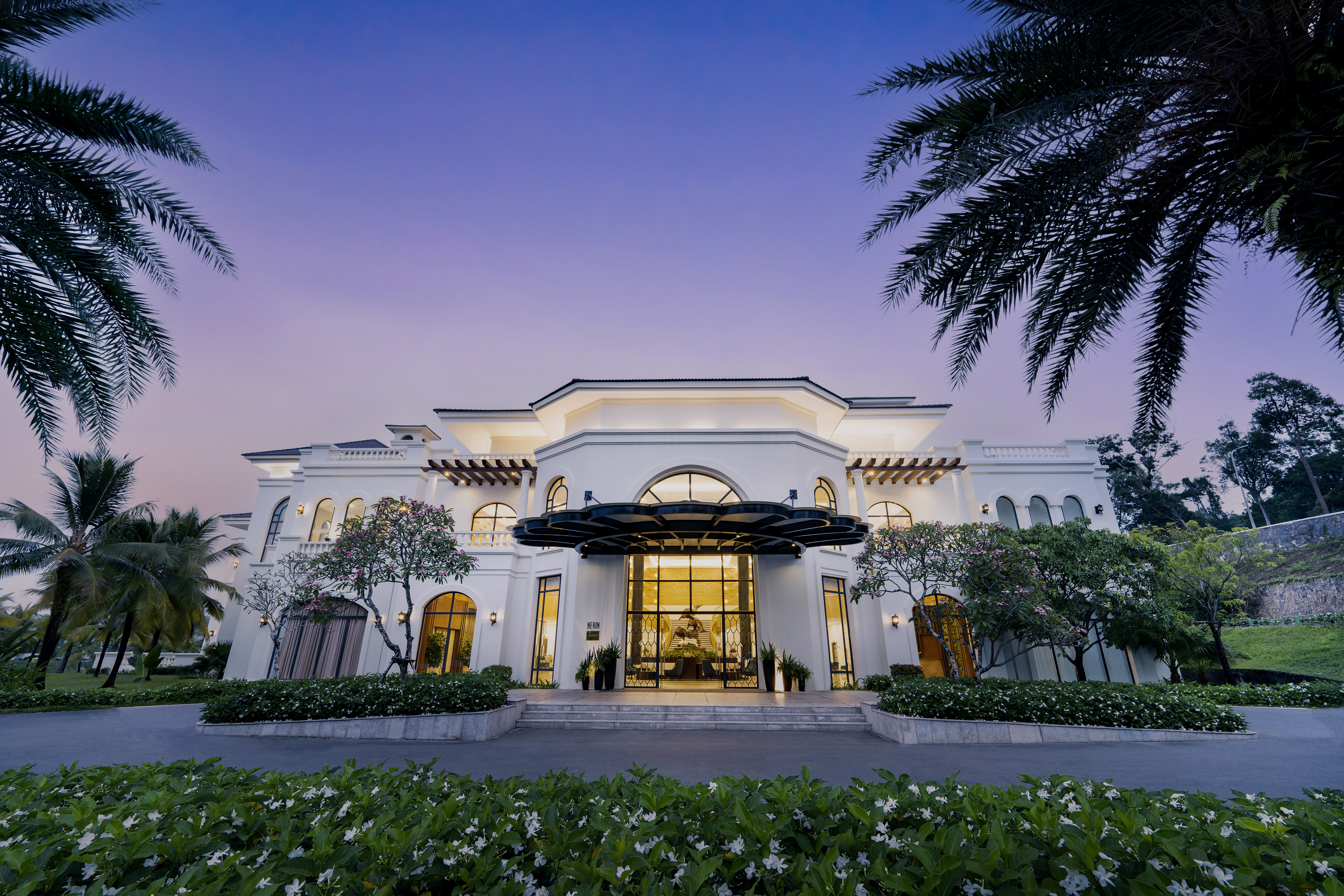 Grand white luxury hotel entrance with palm trees at purple dusk.