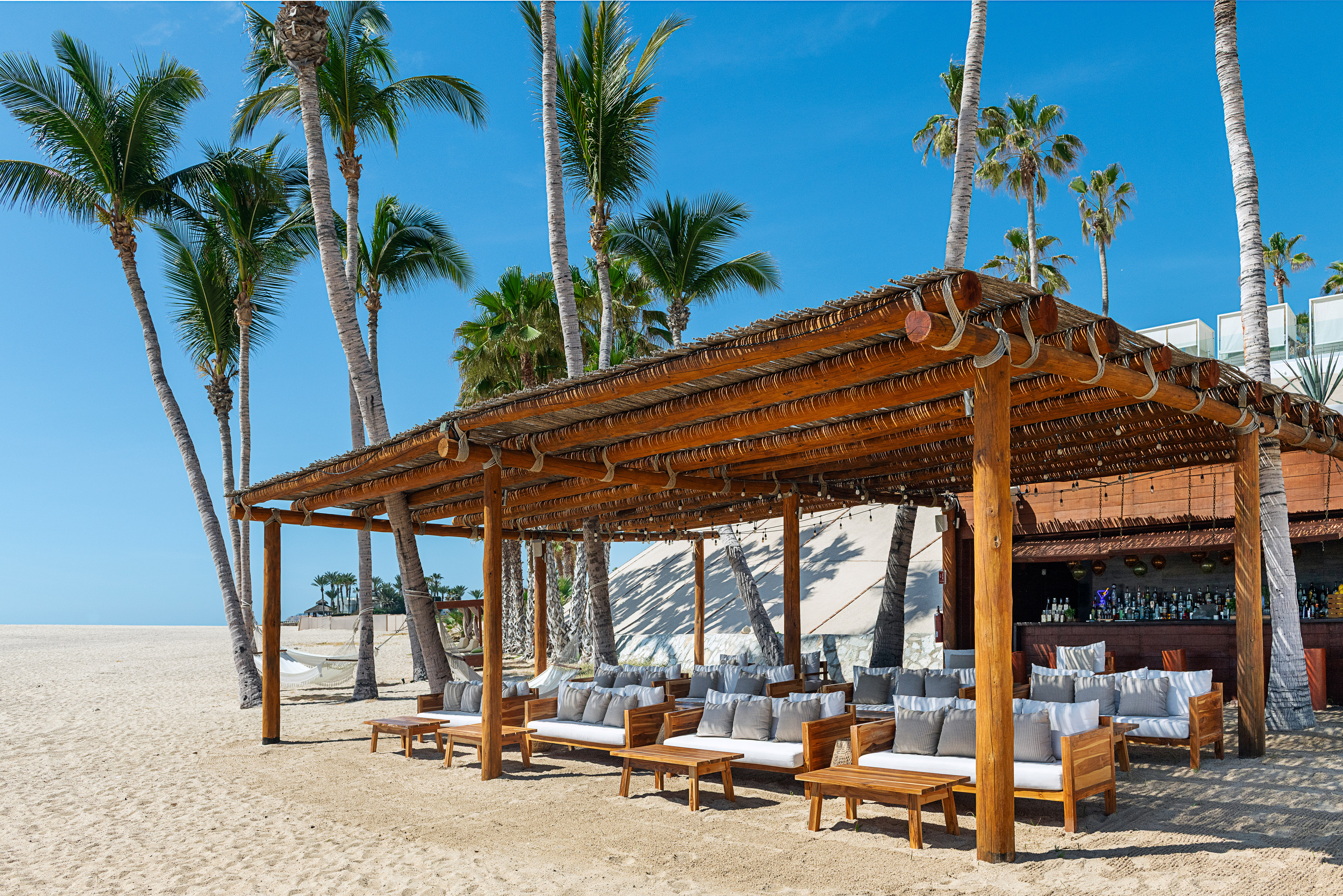Outdoor beach bar and lounge with thatched roof, palm trees, and sandy beach.