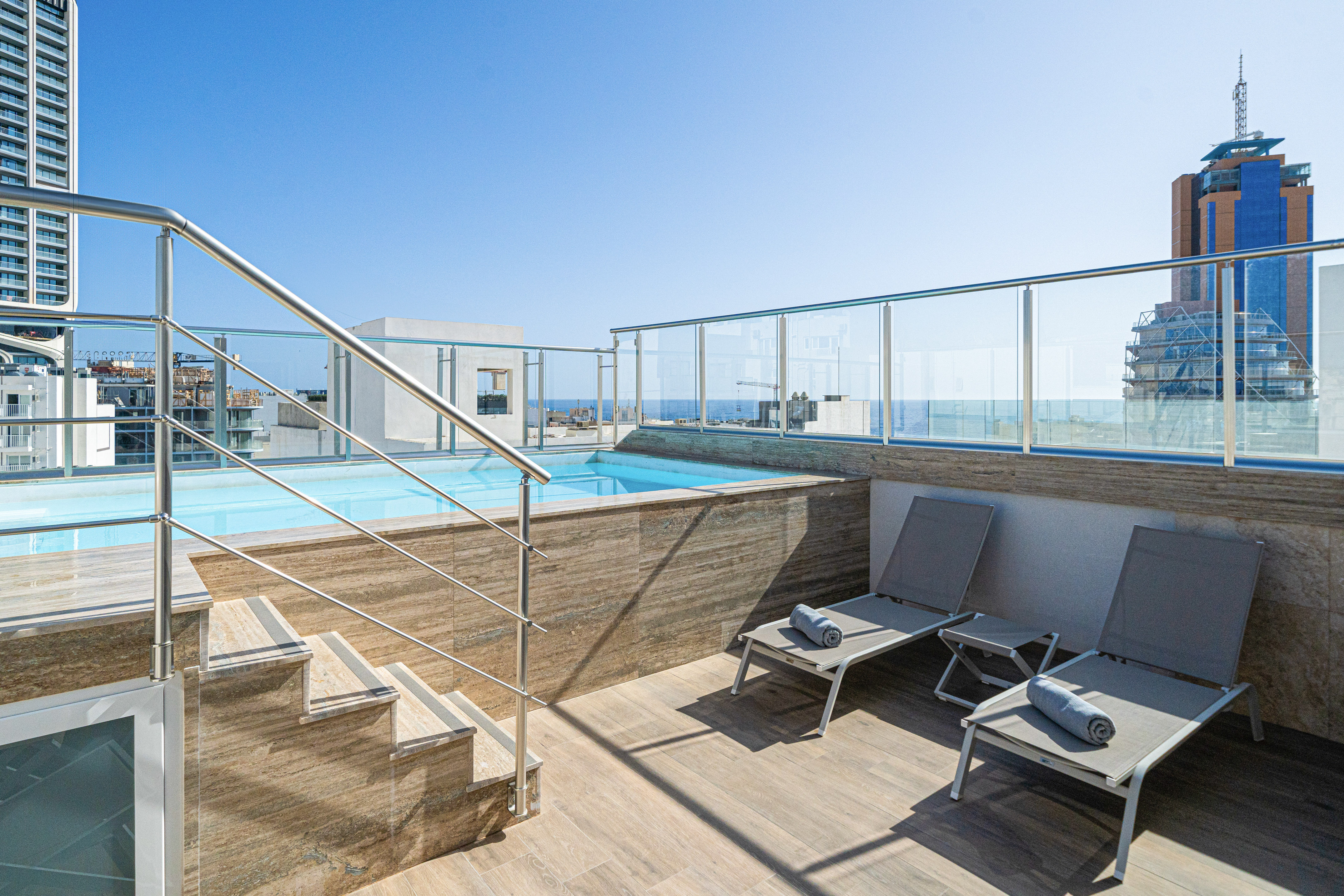 Modern rooftop pool with lounge chairs, glass railing, city and sea views.