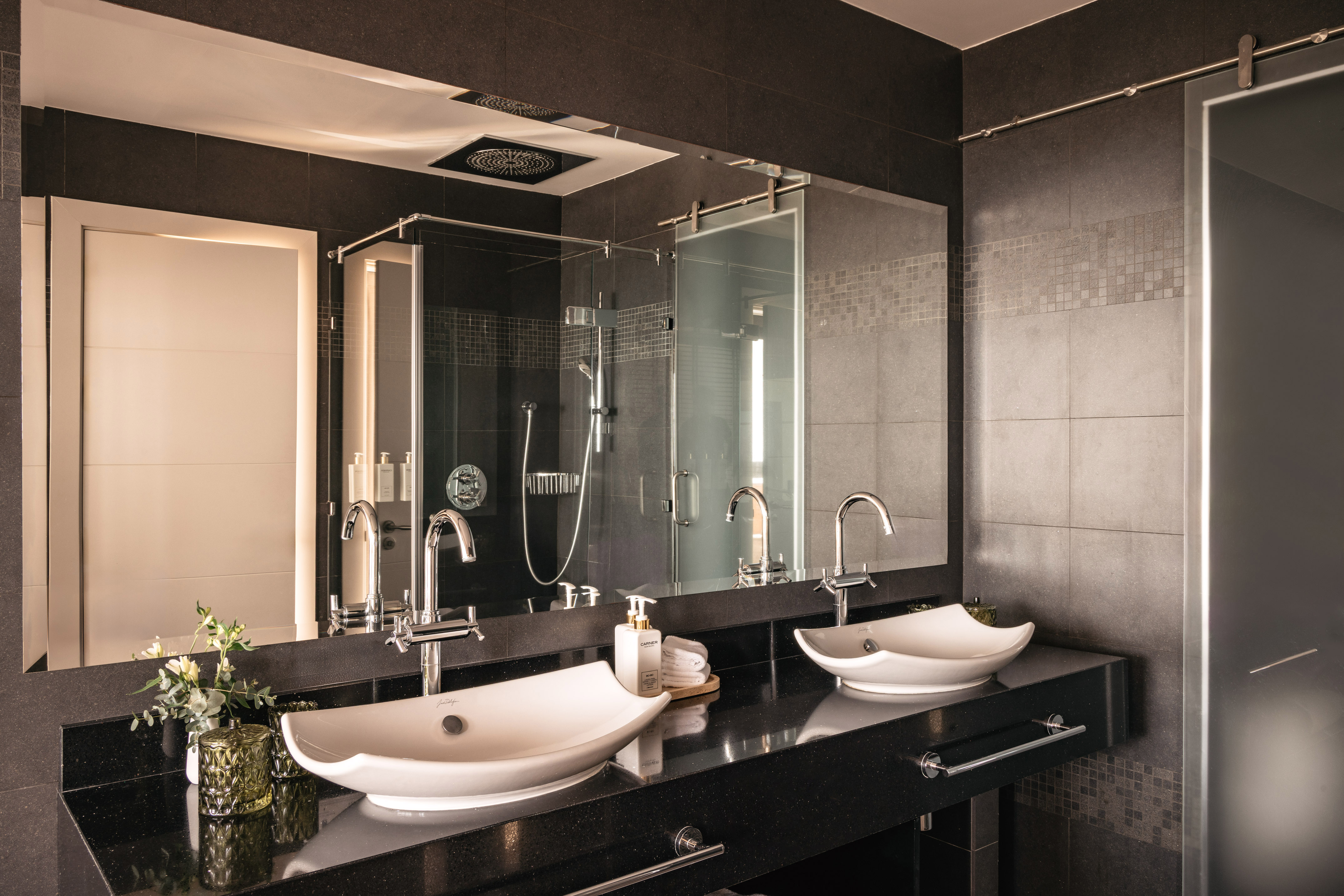 Modern luxury bathroom with double vessel sinks, black countertop, glass shower.