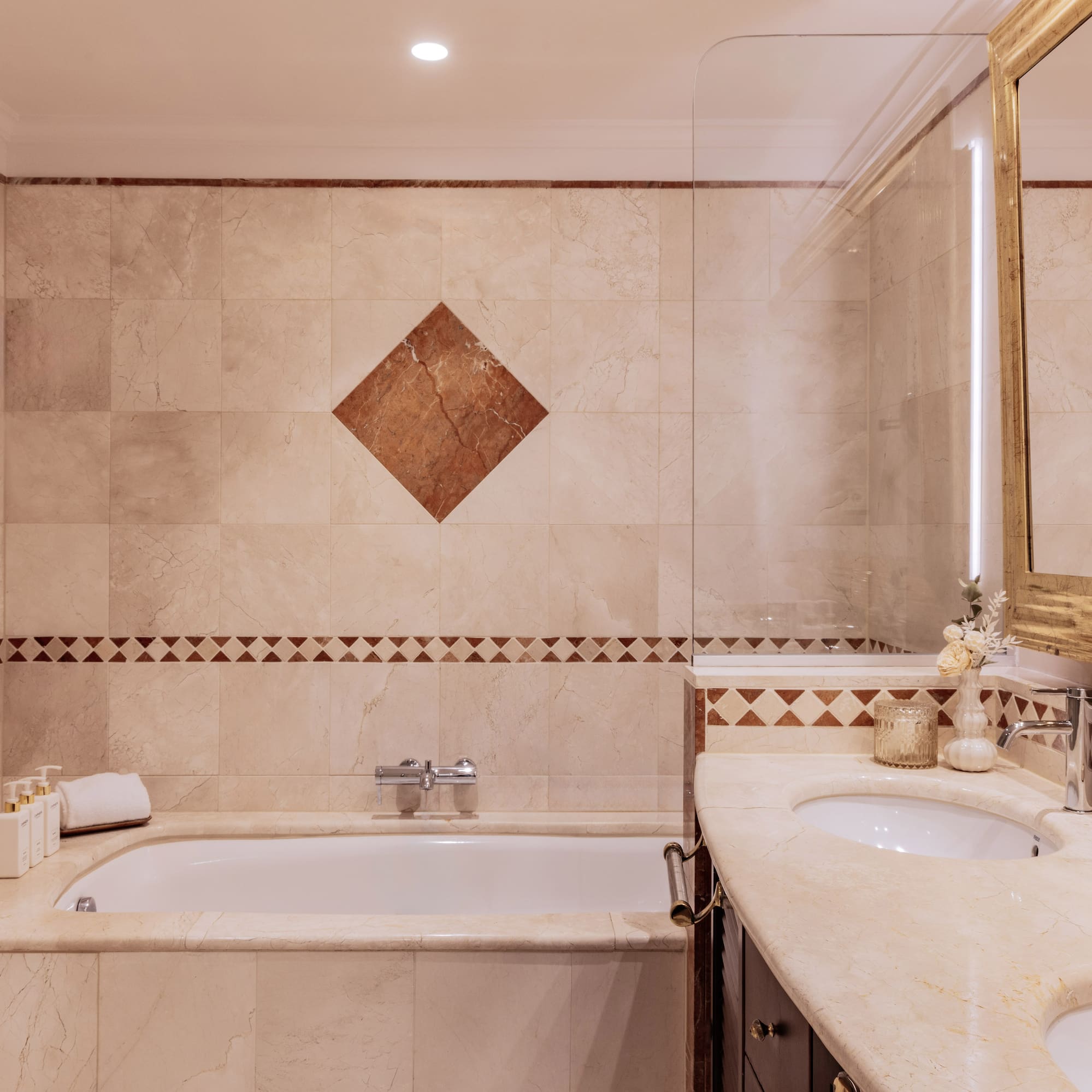 Classic marble bathroom with bathtub, dual sinks, and illuminated golden mirror.