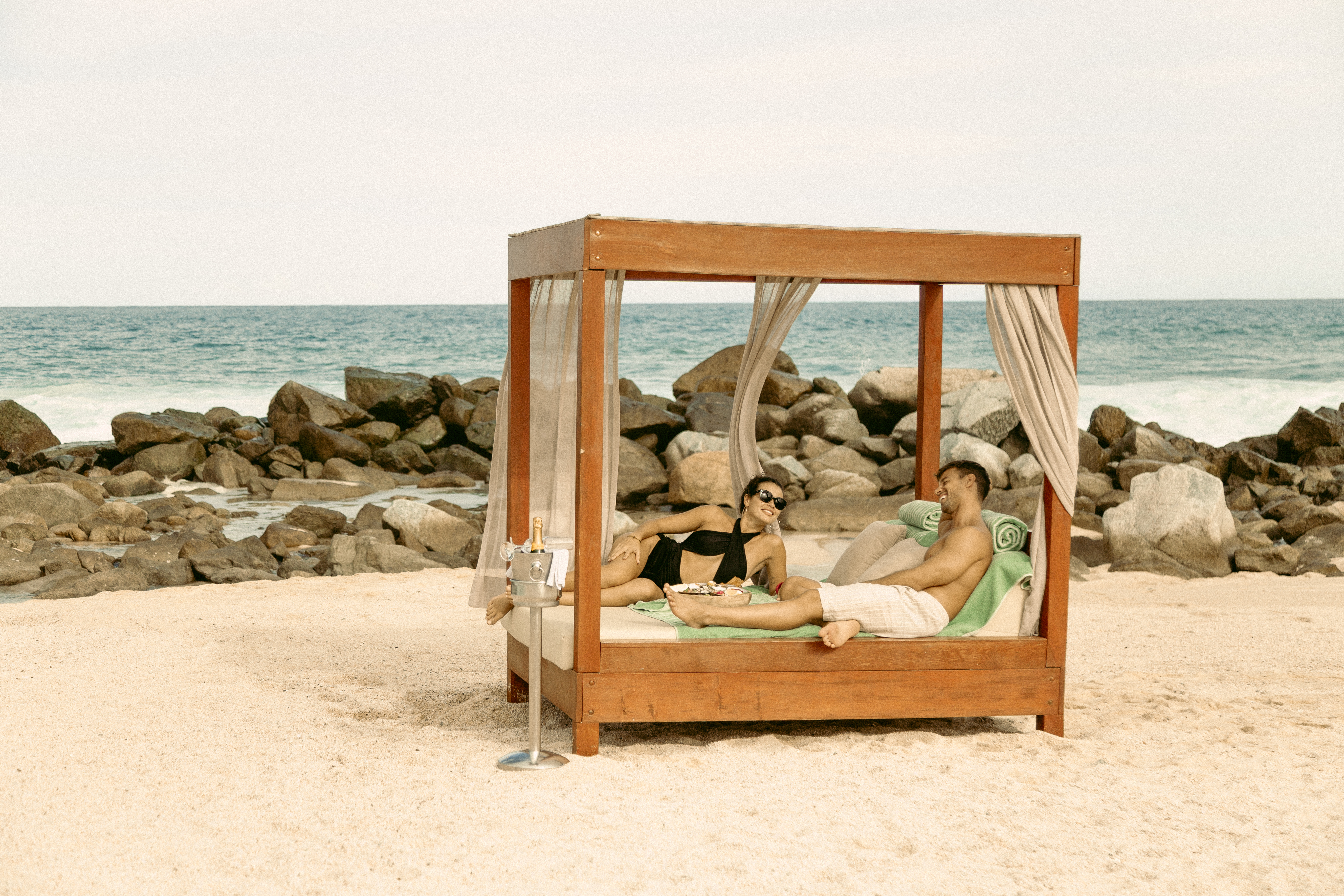 Couple relaxing on a beach cabana bed with champagne by the ocean.