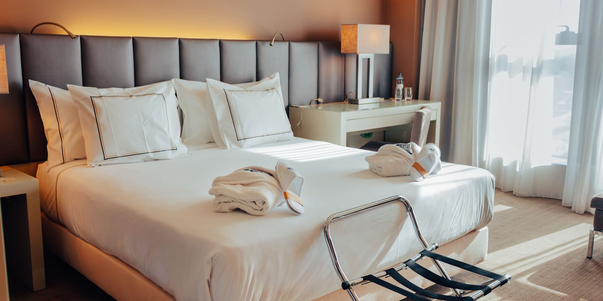 Hotel room with bed, white linens, folded bathrobes, slippers, and luggage rack.