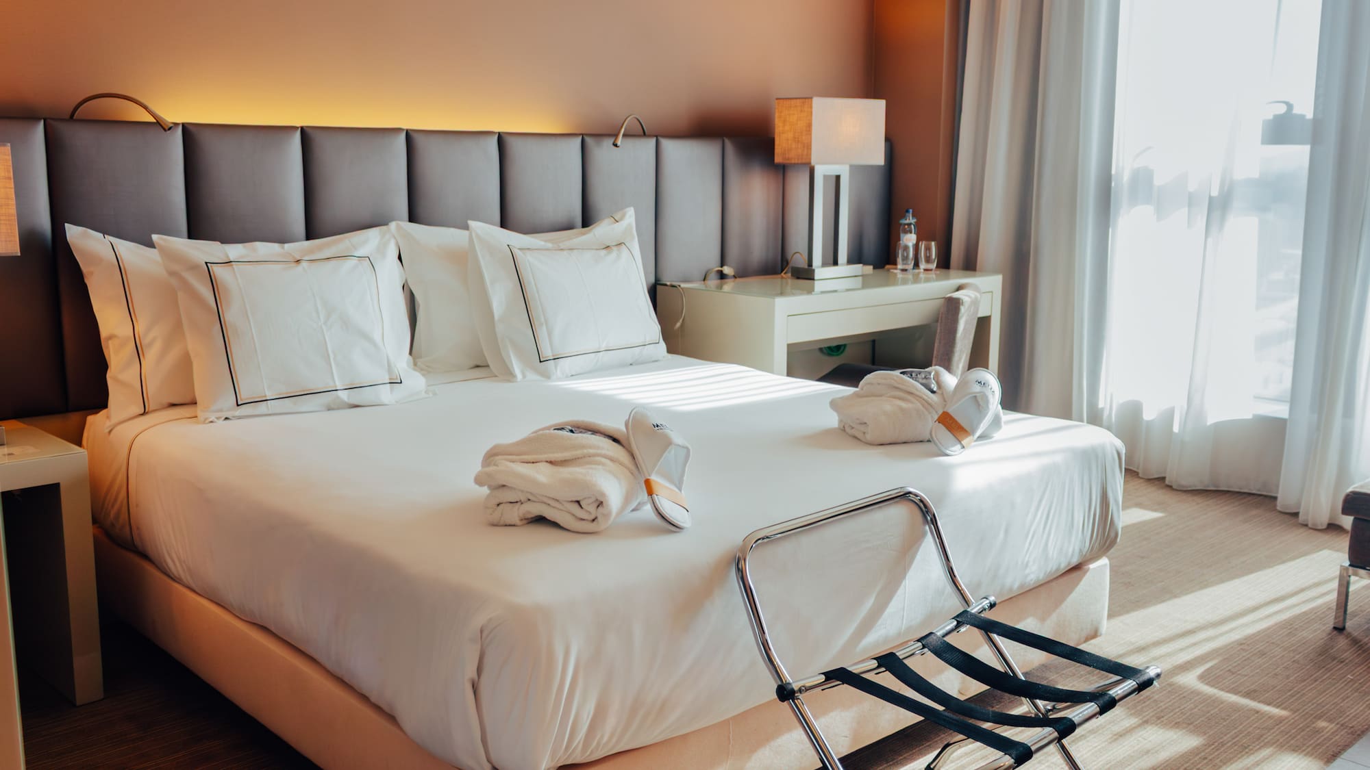 Hotel room with bed, white linens, folded bathrobes, slippers, and luggage rack.