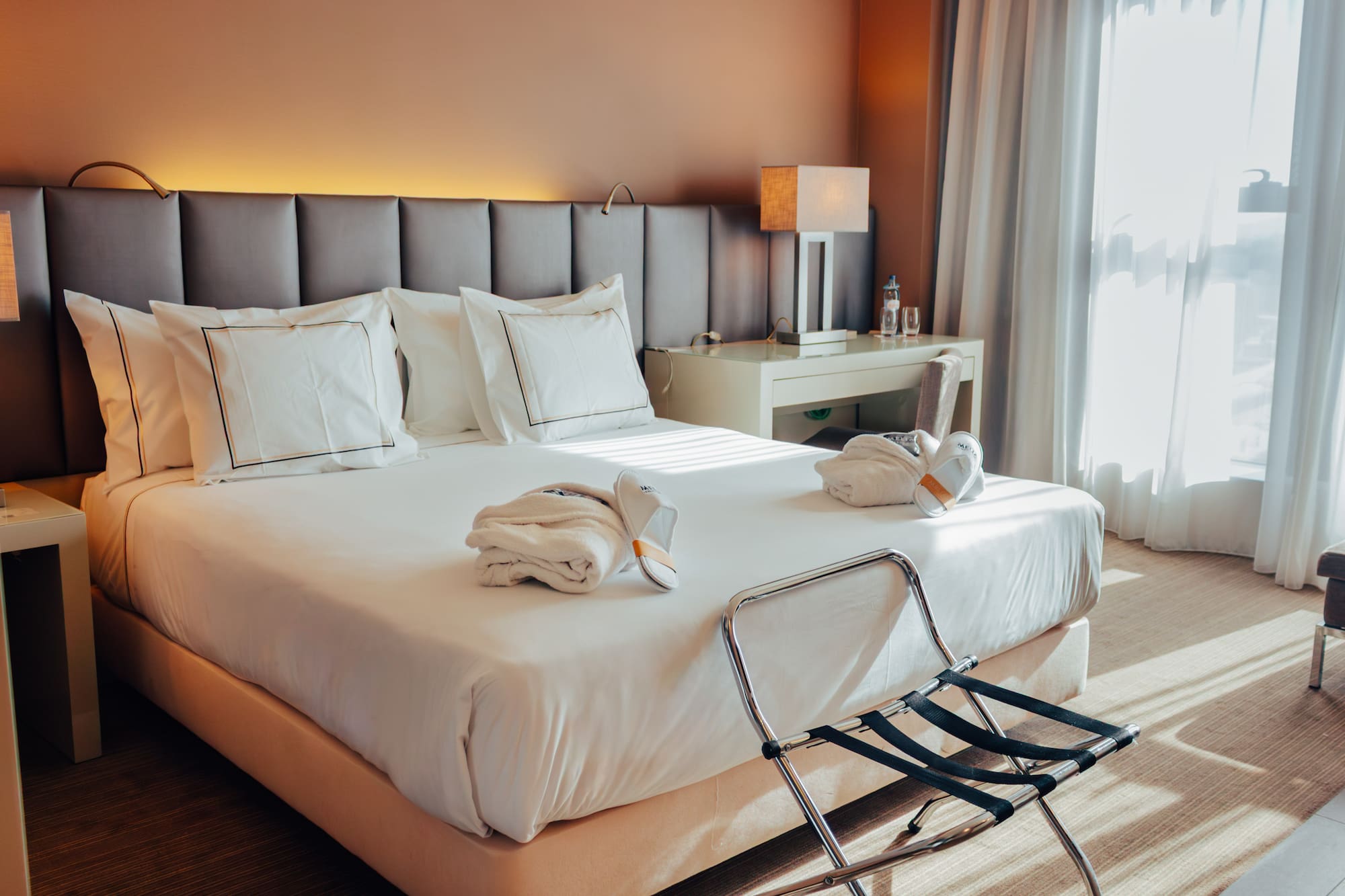 Hotel room with bed, white linens, folded bathrobes, slippers, and luggage rack.
