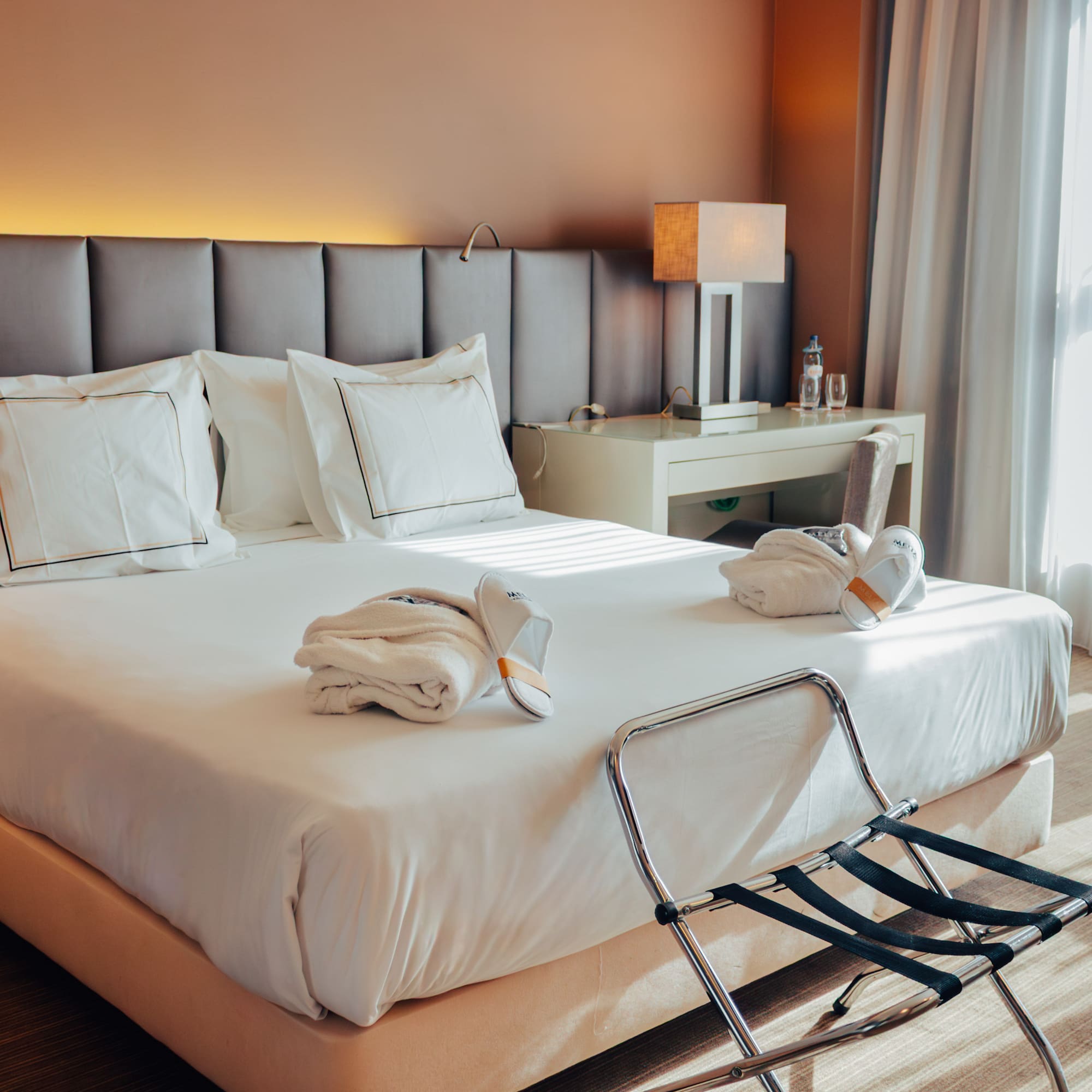Hotel room with bed, white linens, folded bathrobes, slippers, and luggage rack.