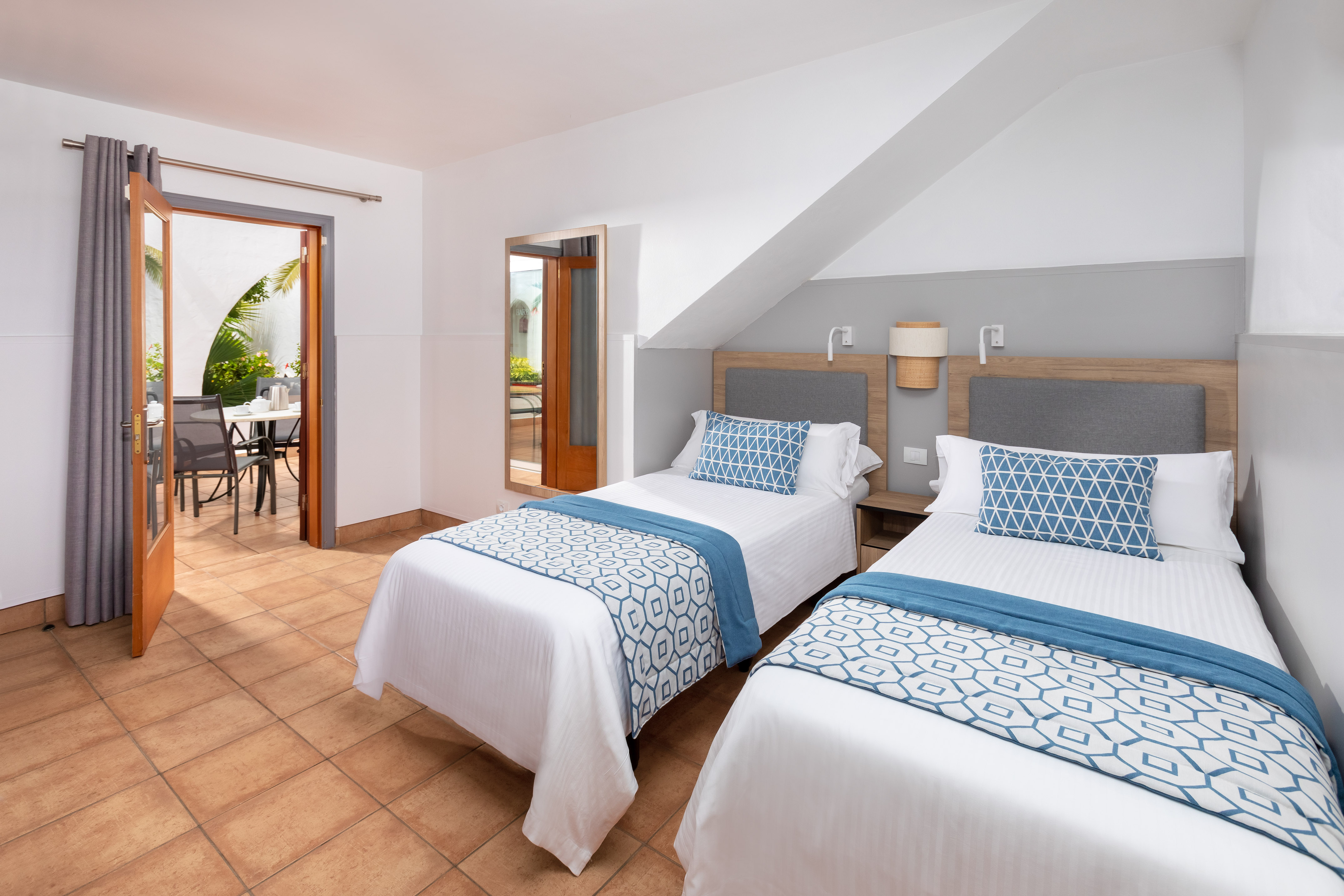 Hotel twin room, two single beds with blue accents, tiled floor, patio view.