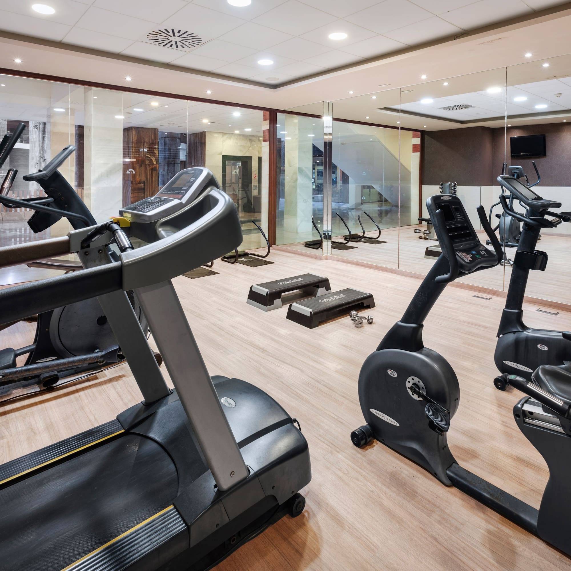 Fitness room with treadmill, elliptical, stationary bikes, and dumbbells on wood floor.