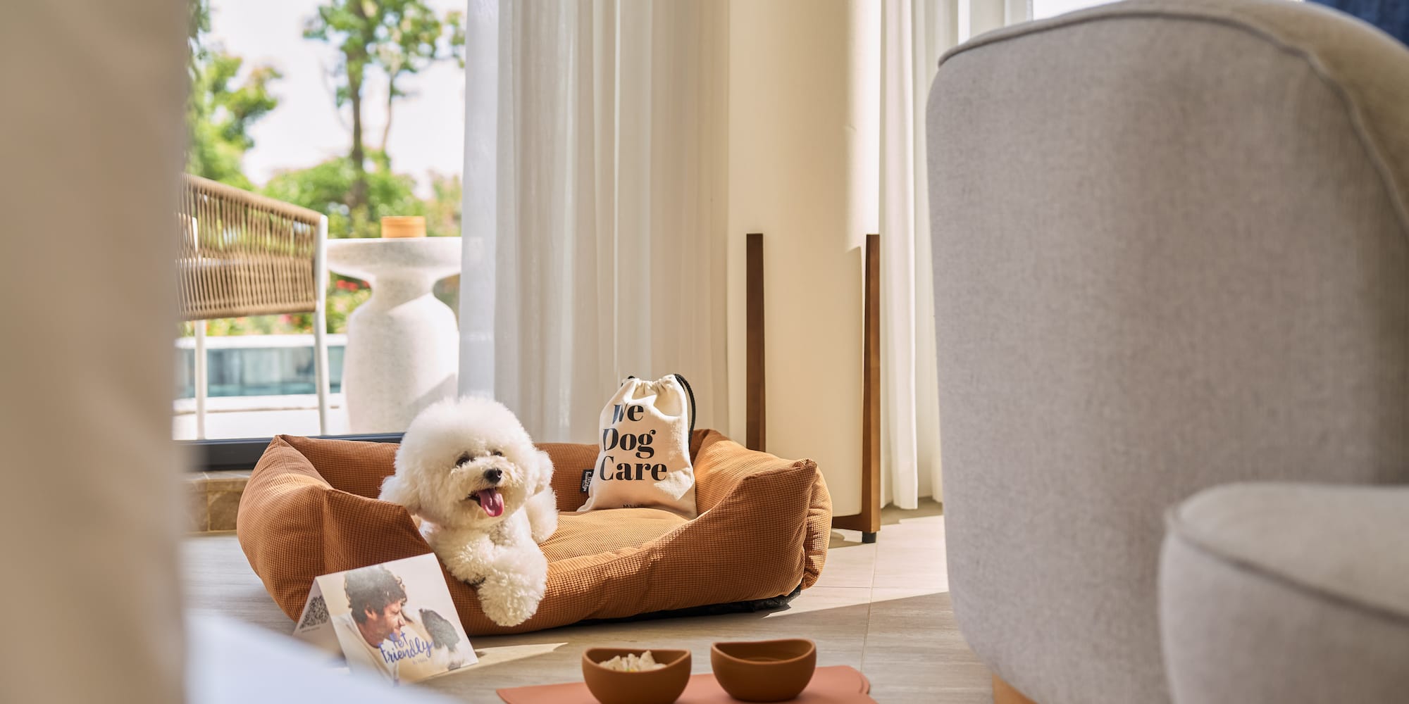 Bichon Frise in pet bed with bowls, "We Dog Care" bag, bright pet-friendly room.