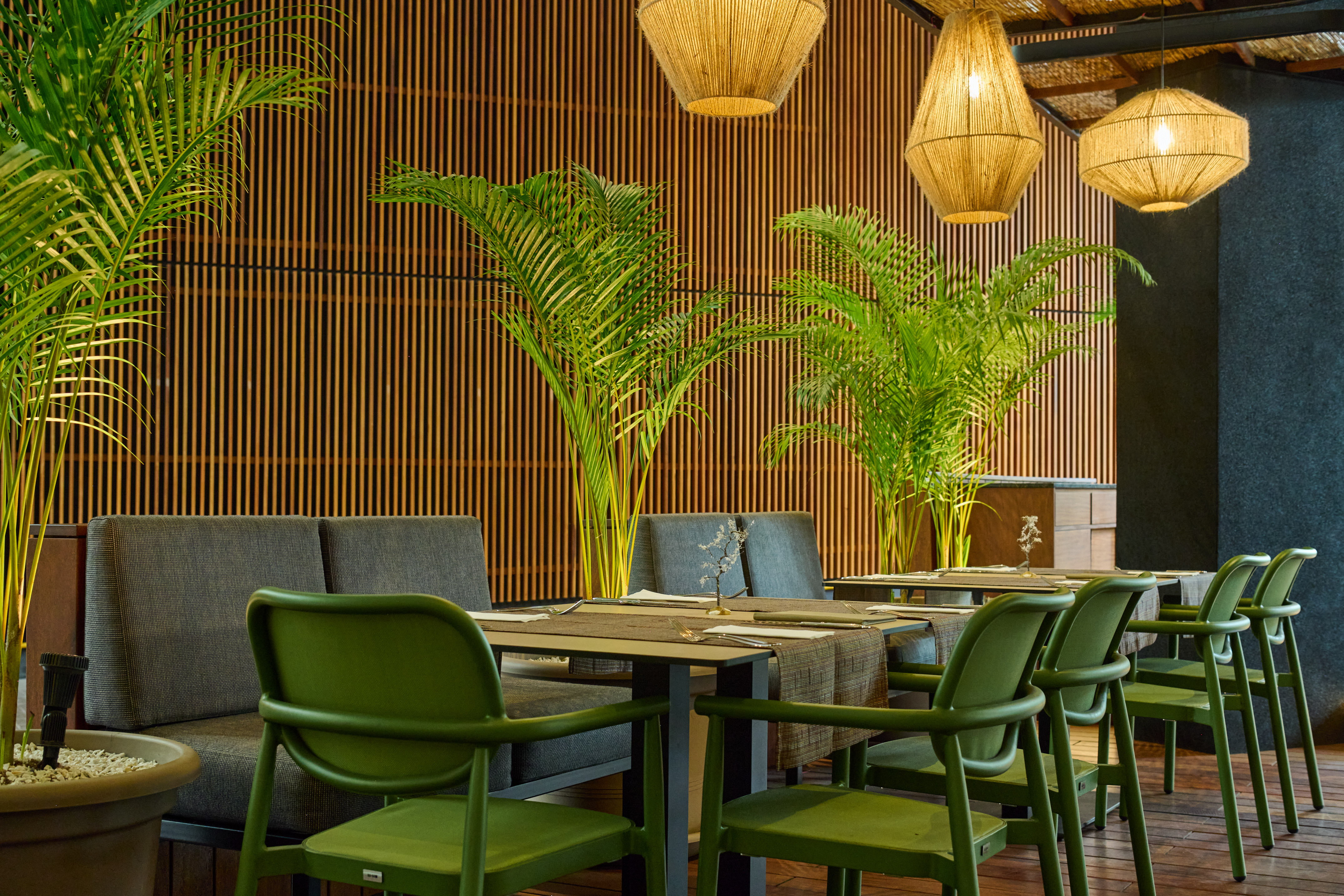 Modern restaurant interior with green chairs, palm plants, rattan lights, wood walls.