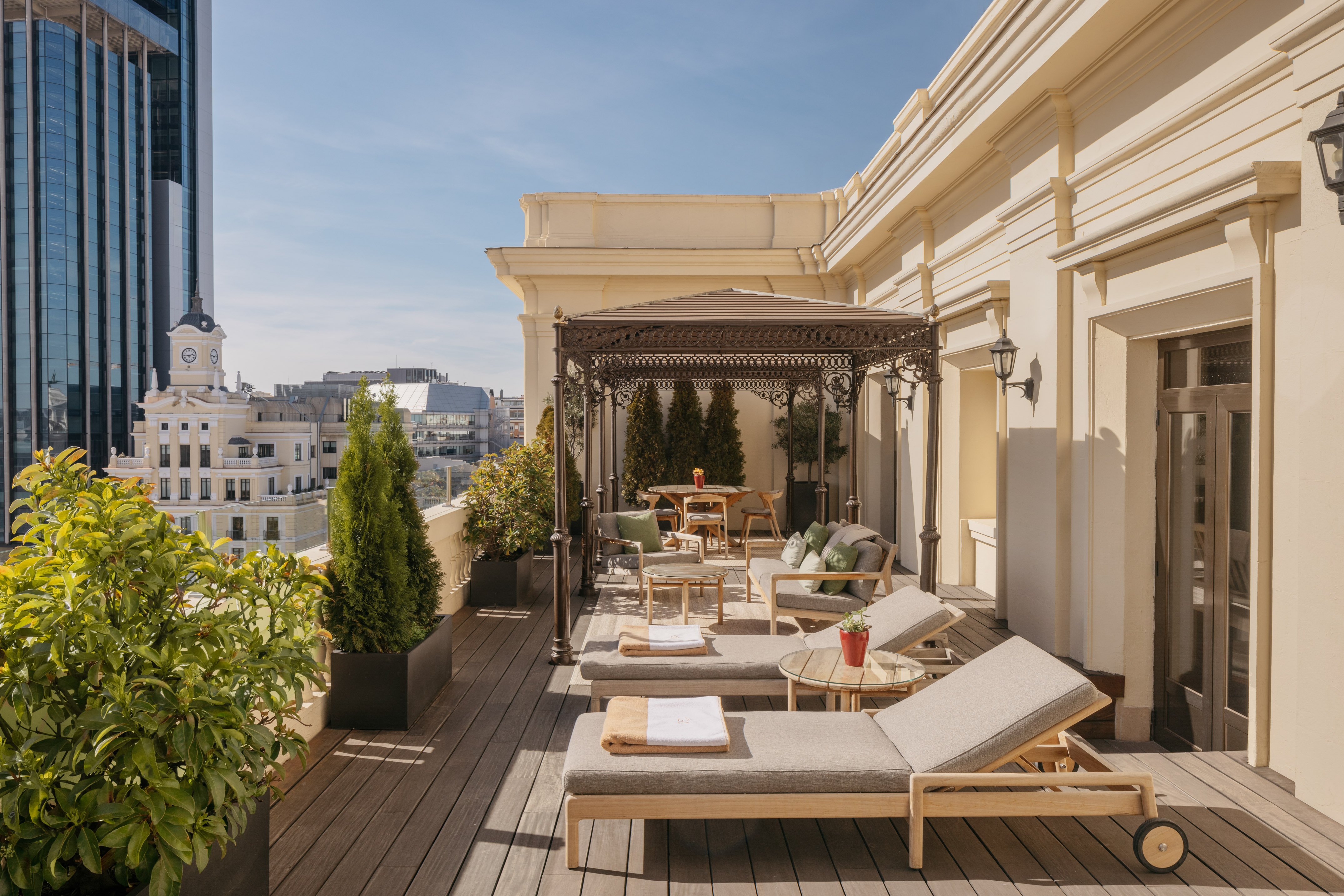 Sunny hotel rooftop terrace with lounge chairs, dining gazebo, and city view.