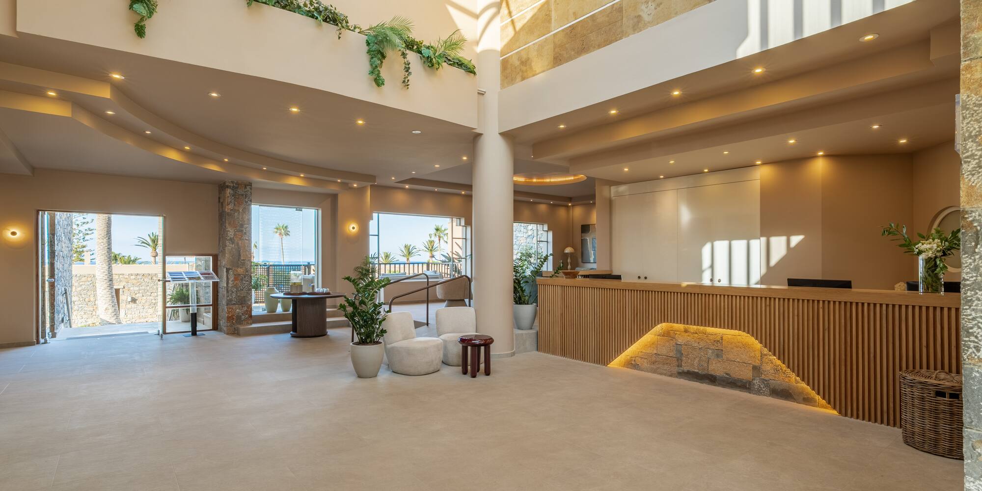Bright hotel lobby with sea view, modern reception desk, and lush plants.