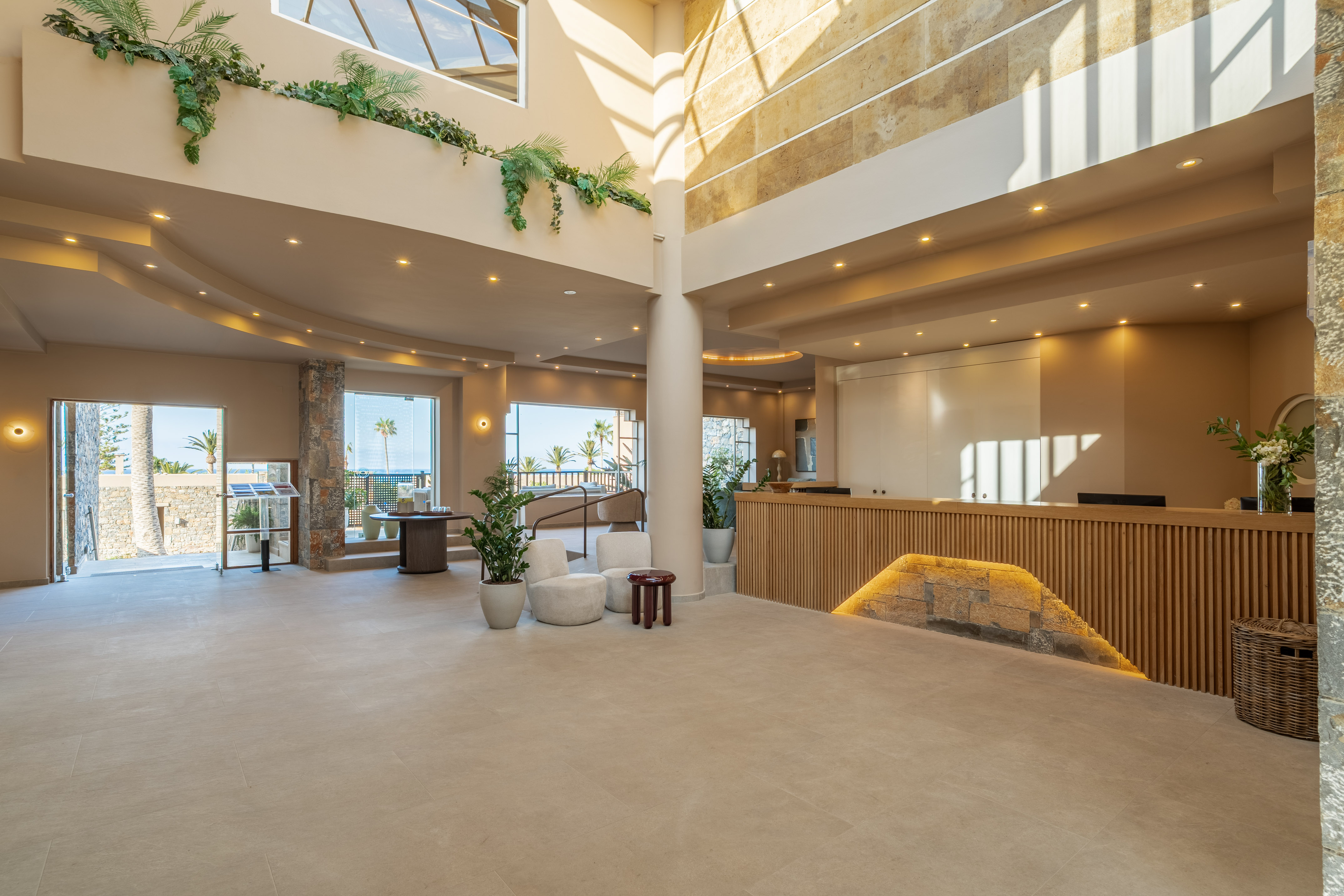 Bright hotel lobby with sea view, modern reception desk, and lush plants.