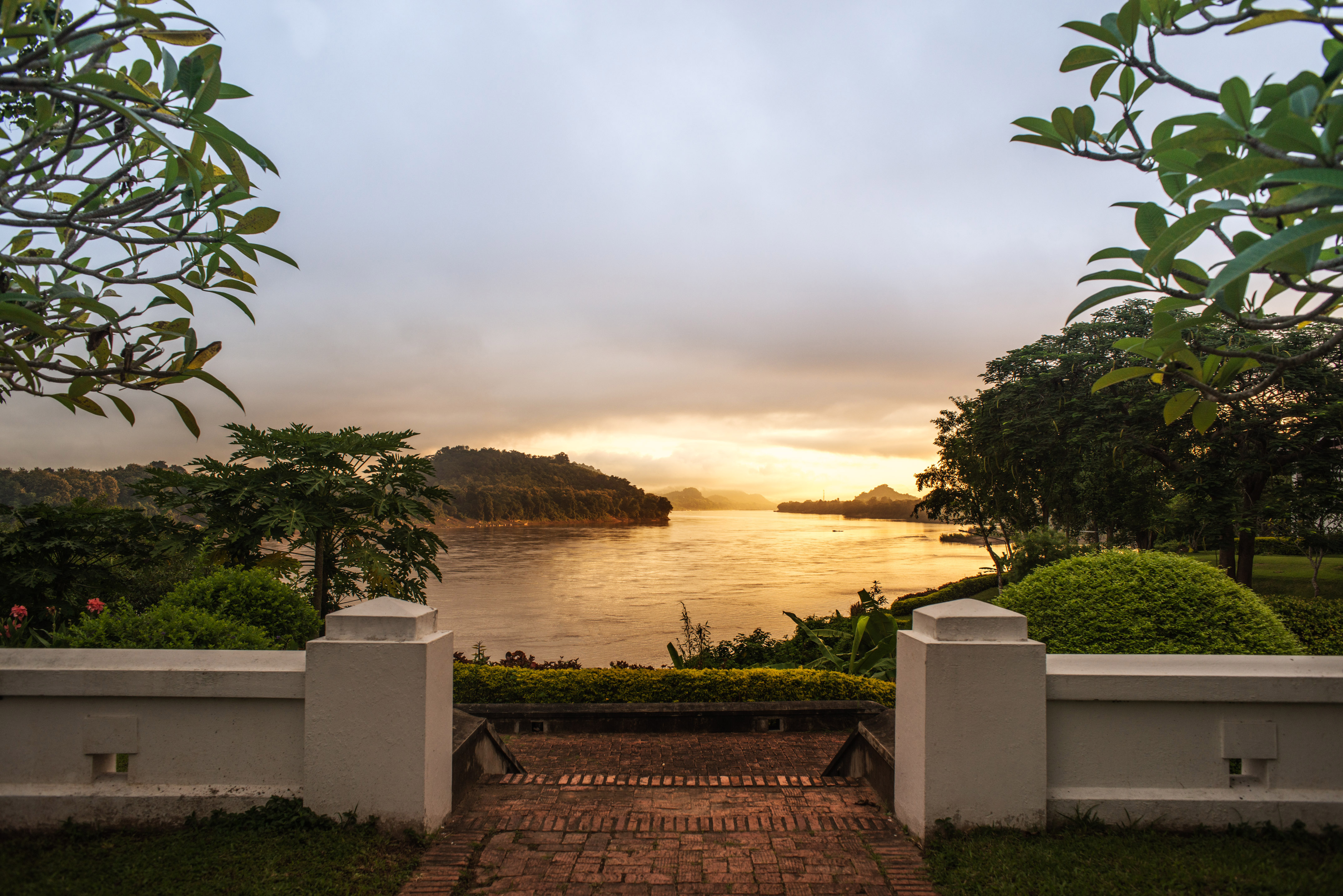 Brick path leading to golden river sunset framed by lush tropical trees.