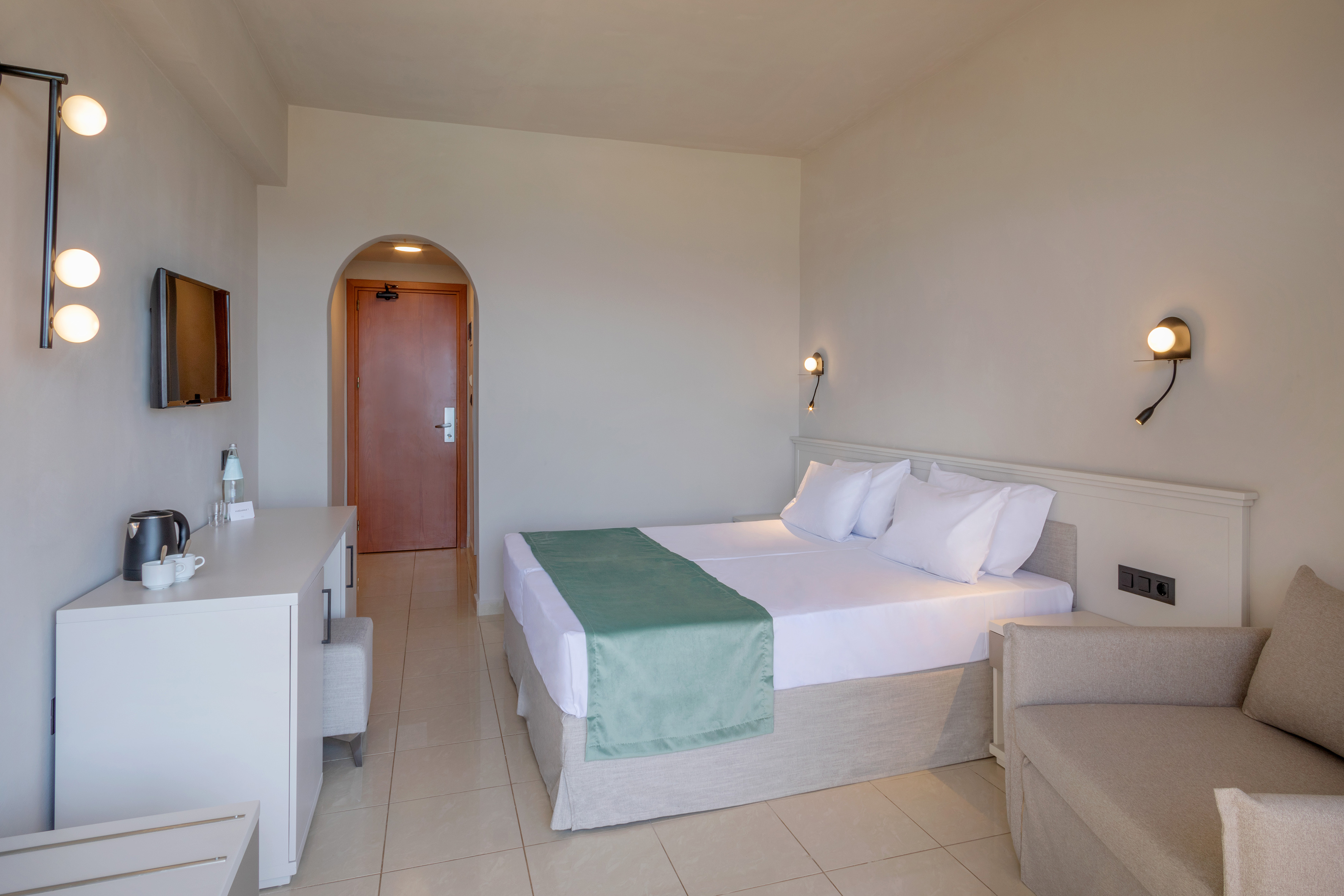 Modern hotel room with twin beds, green bed runner, TV, and sofa.