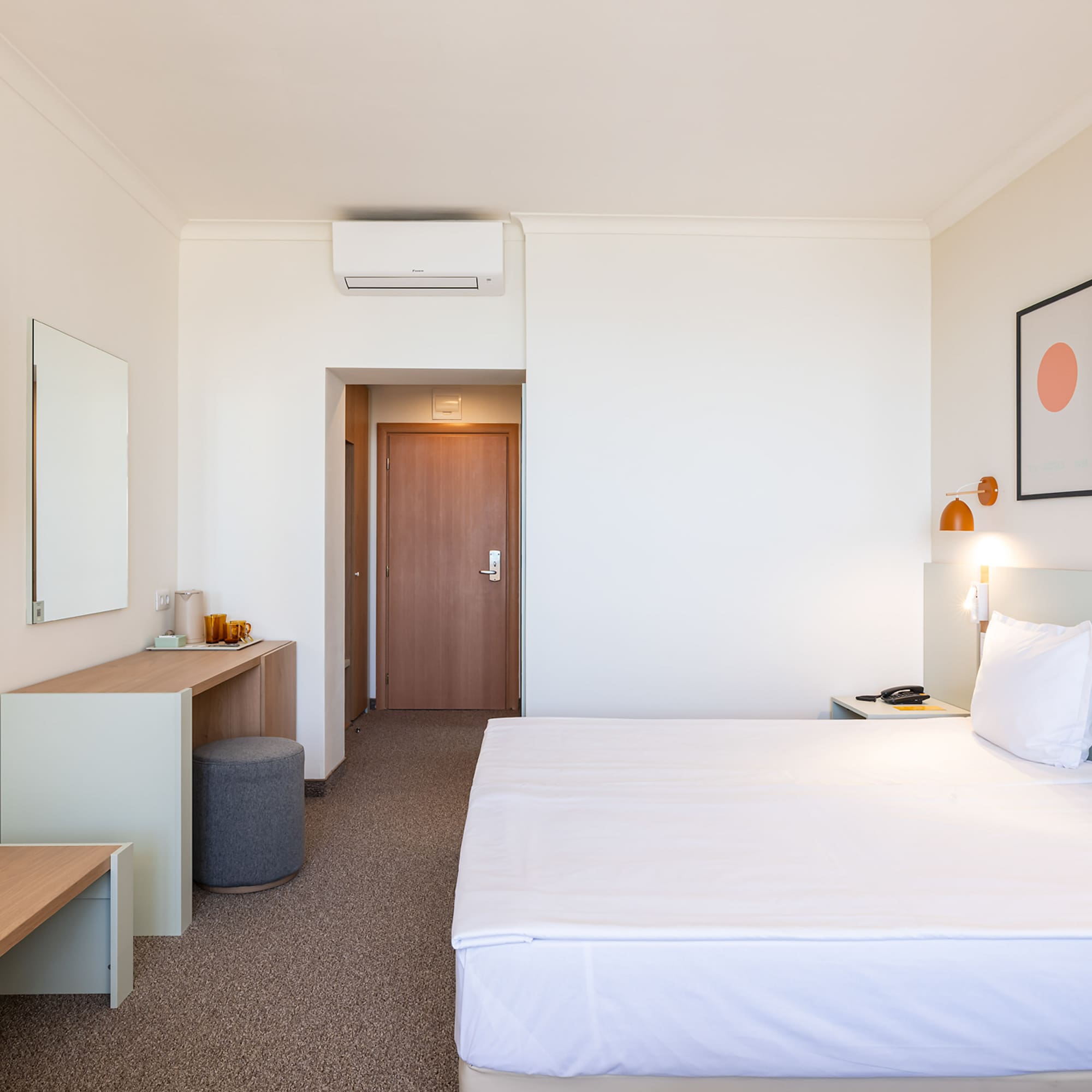 Modern hotel bedroom with white bed, desk, wall-mounted TV, and framed art.