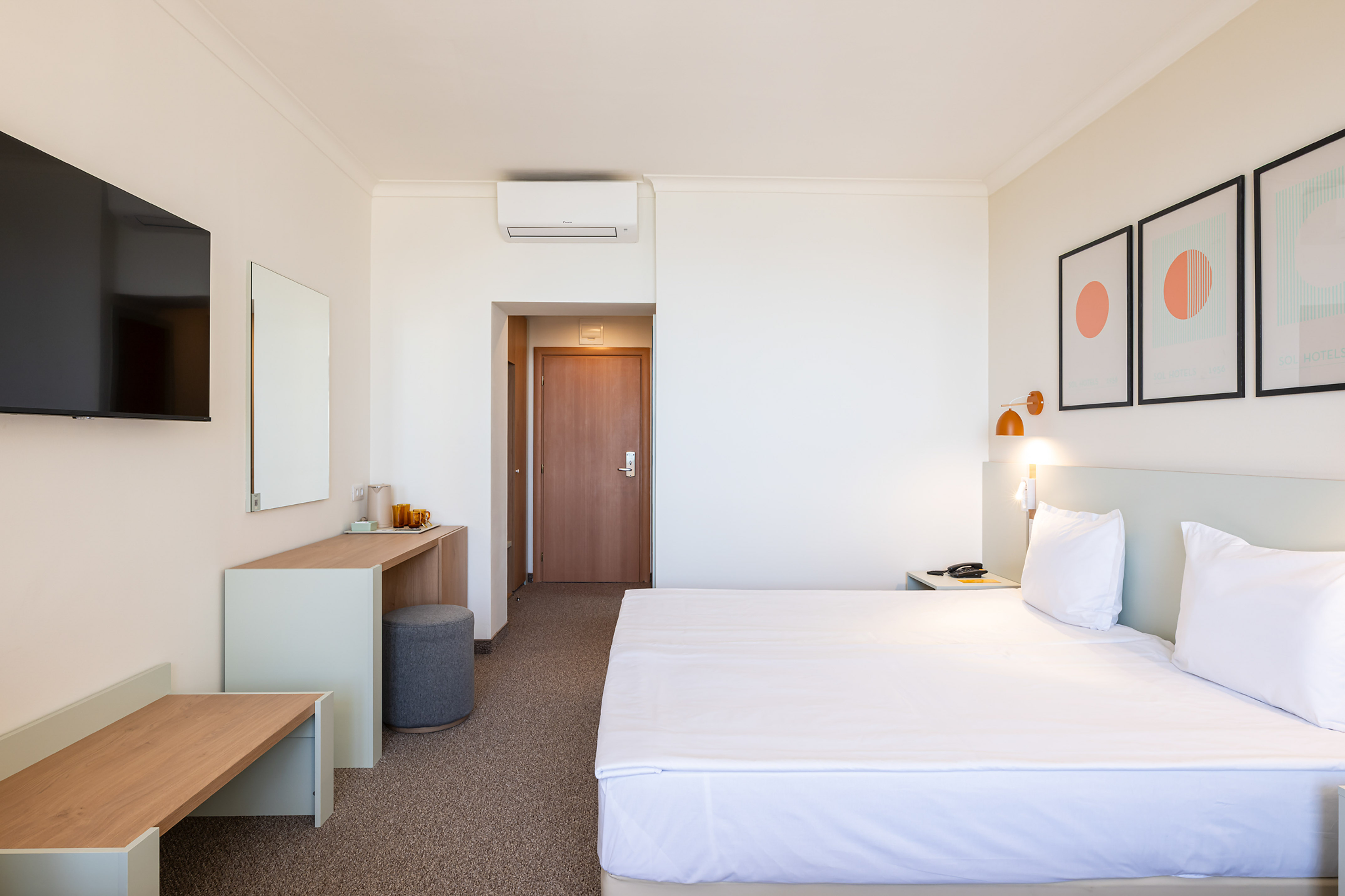 Modern hotel bedroom with white bed, desk, wall-mounted TV, and framed art.
