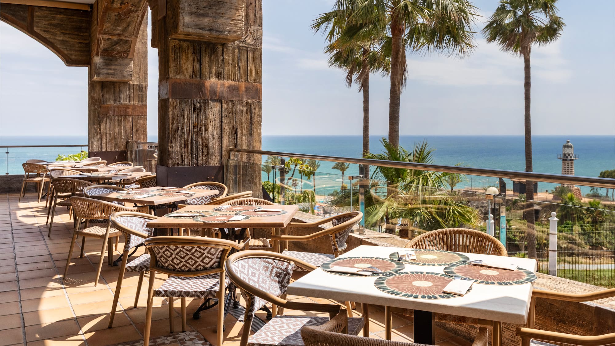 Oceanfront restaurant terrace with palm trees and a distant lighthouse.