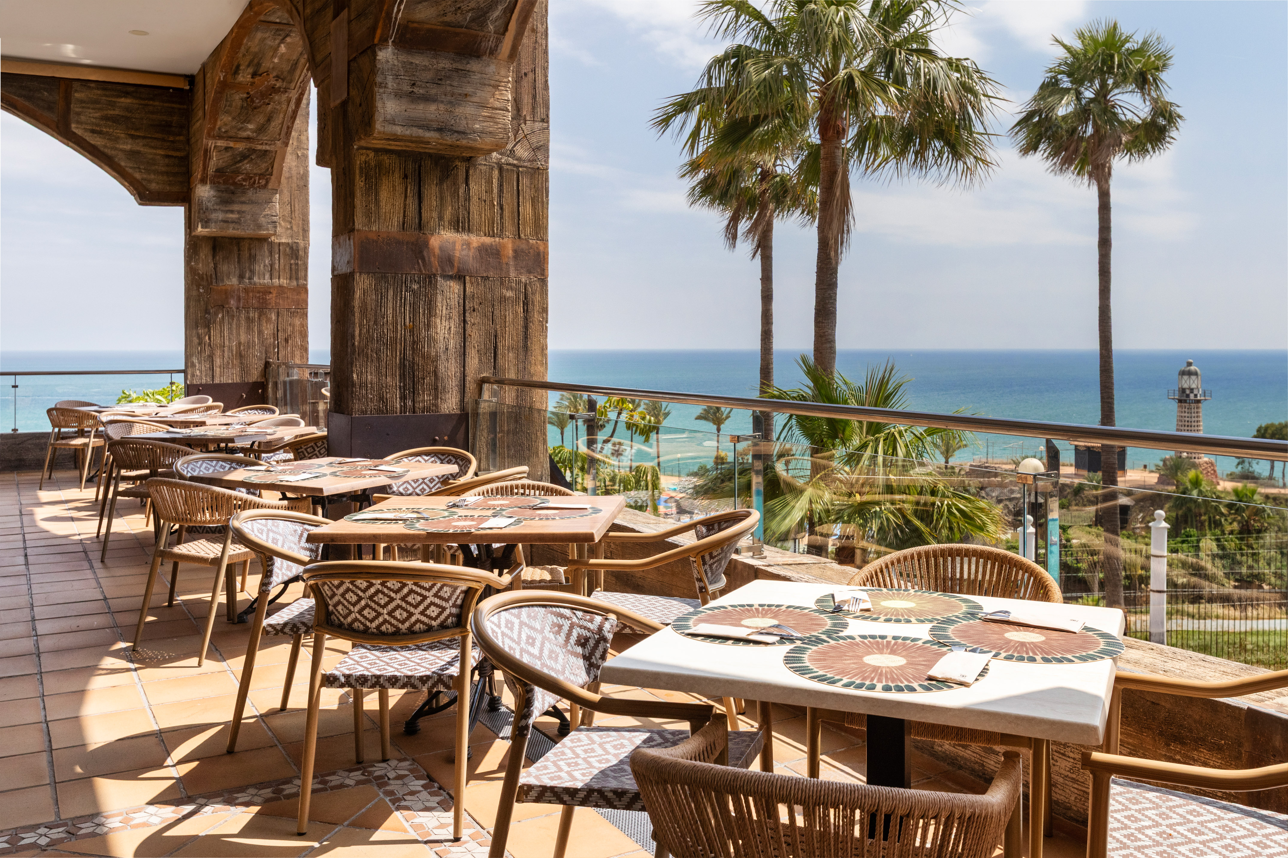 Outdoor ocean view restaurant terrace with palm trees and a distant lighthouse.