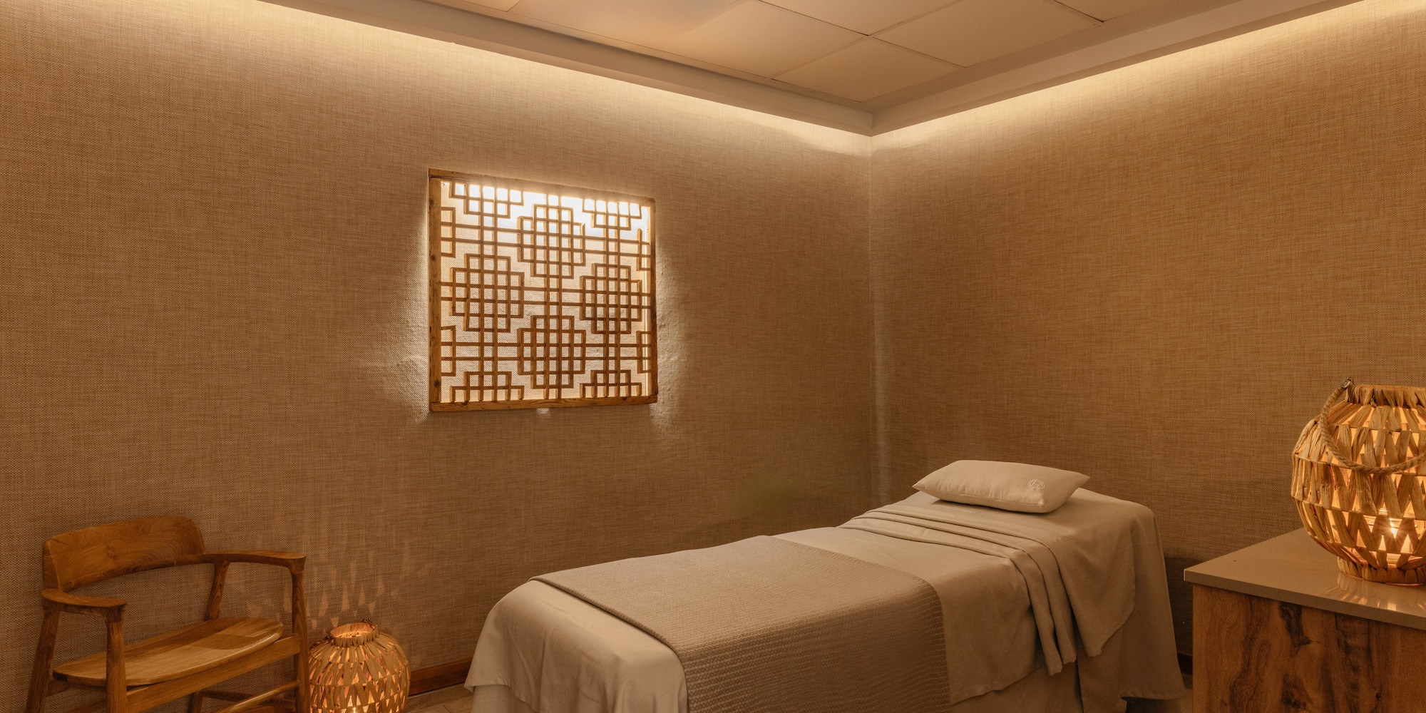 Warm-lit spa room with massage bed, decorative wooden screen, and chair.