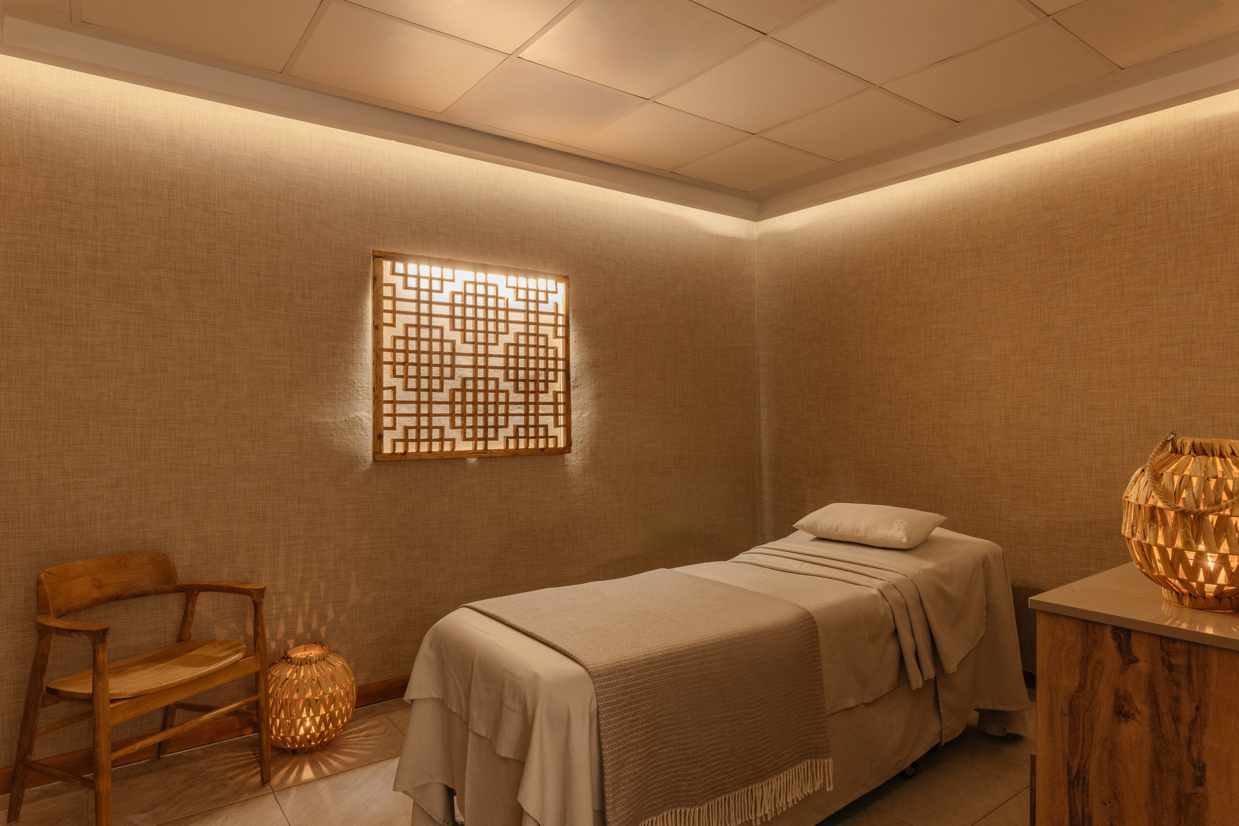 Warm-lit spa room with massage bed, decorative wooden screen, and chair.