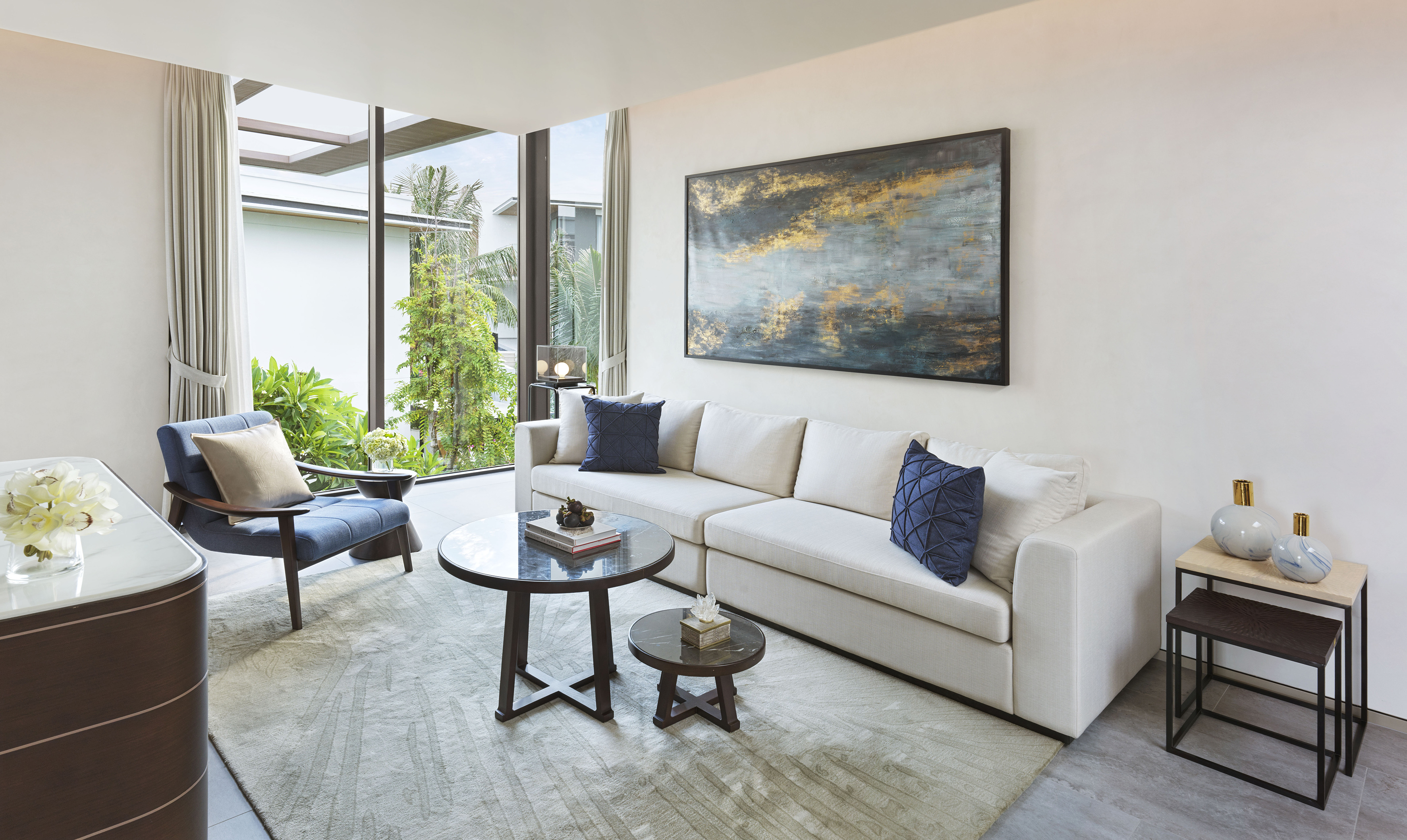 Modern living room with white sofa, blue armchair, abstract art, and garden views.