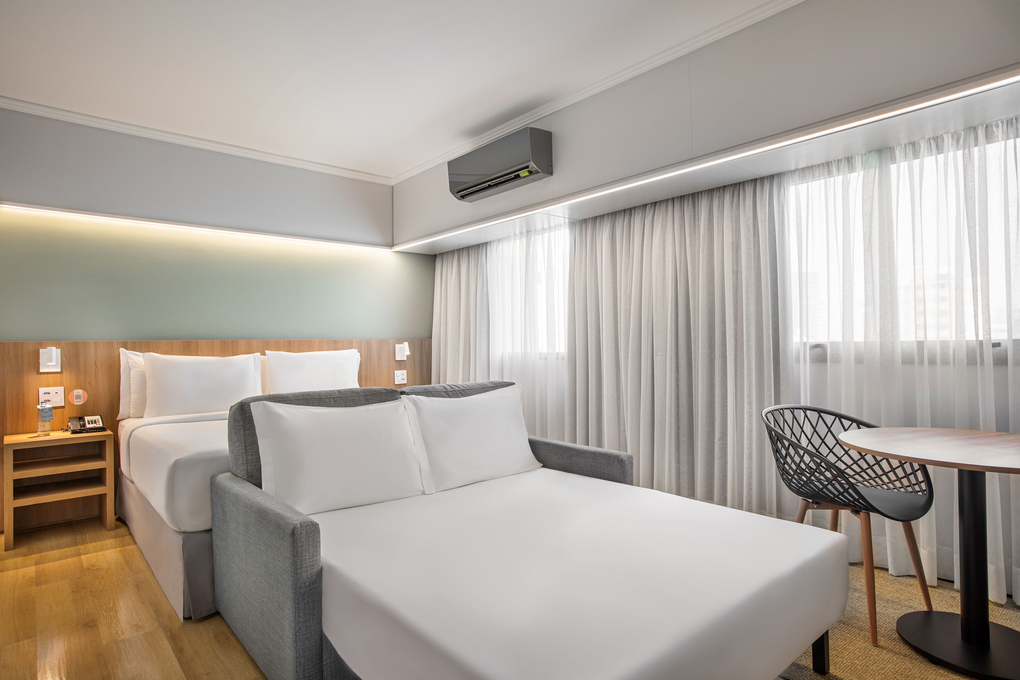 Modern hotel room with bed, sofa bed, desk, and large window with curtains.