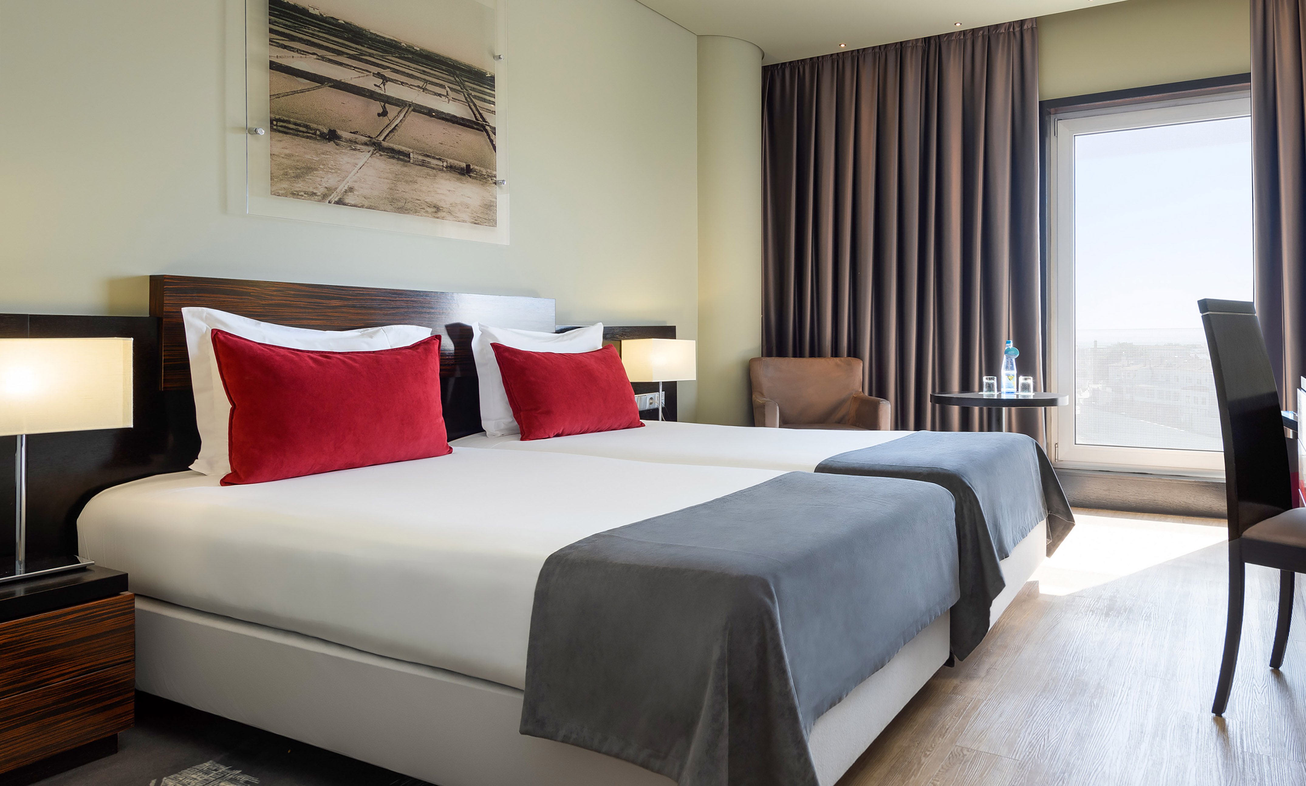 Bright hotel room with two single beds, red pillows, and large window.