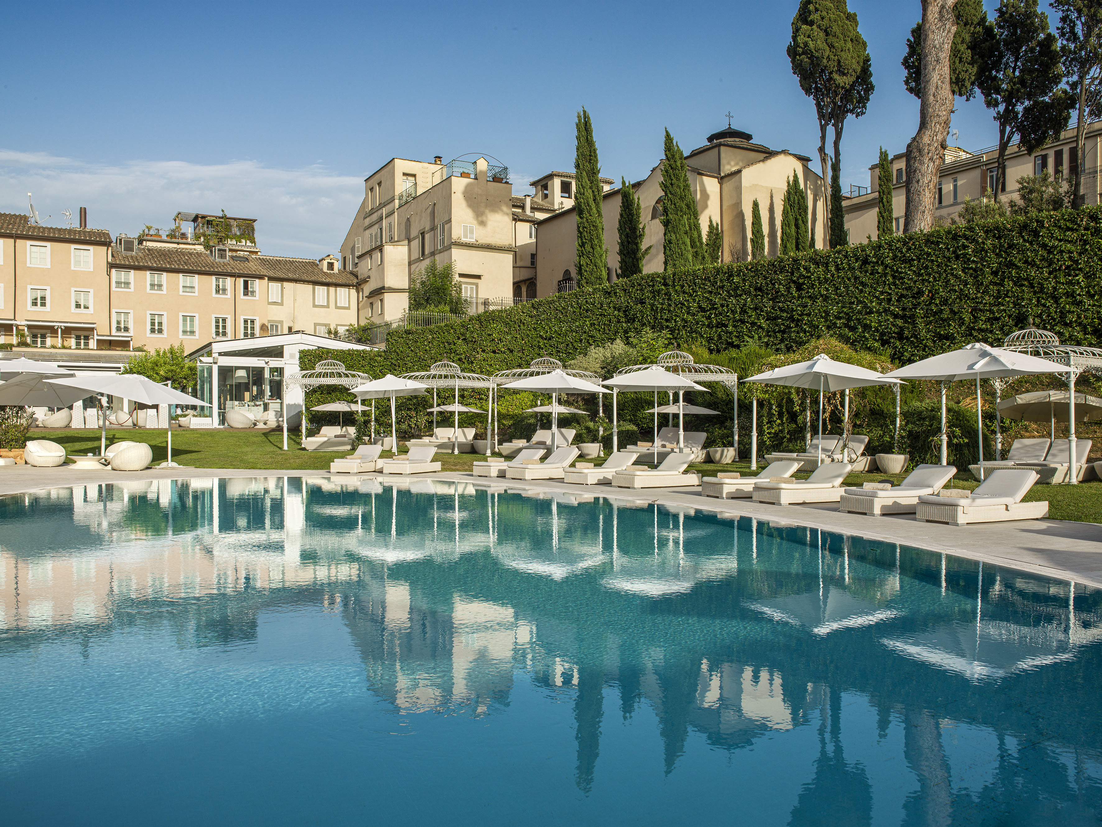 Luxury hotel swimming pool, white loungers, umbrellas, historic buildings, trees.