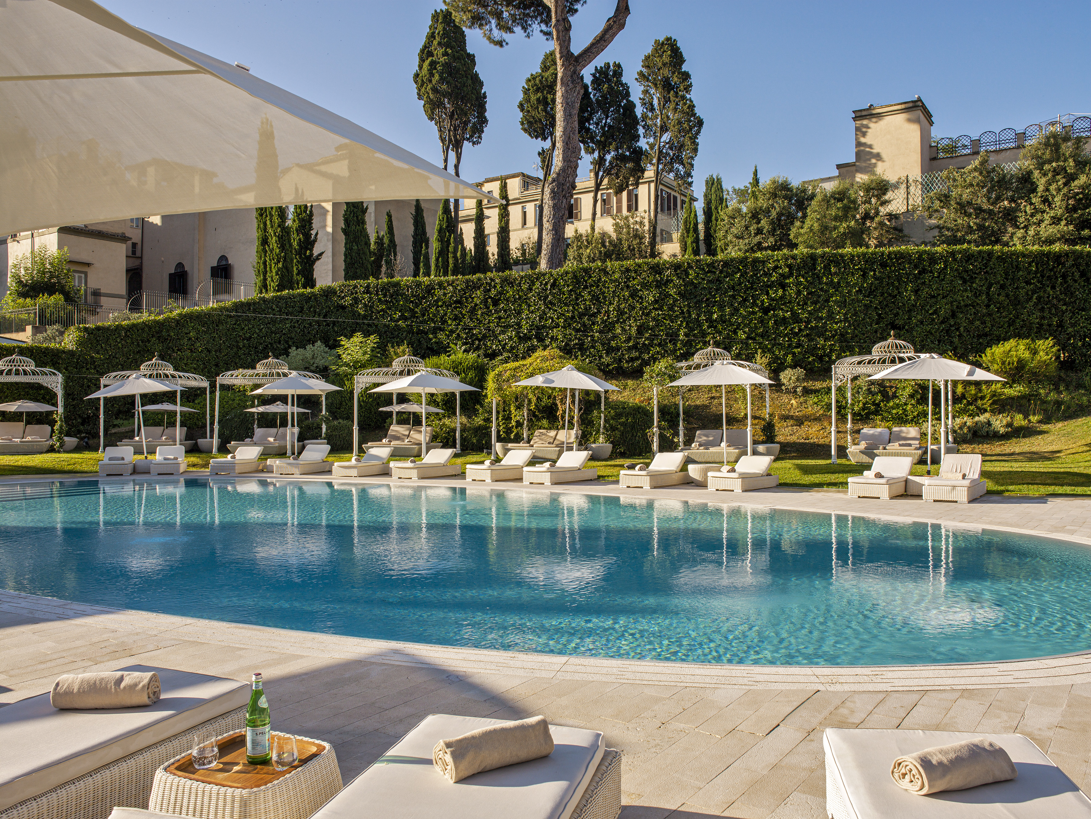 Luxury resort pool with white sunbeds, umbrellas, and historic buildings.