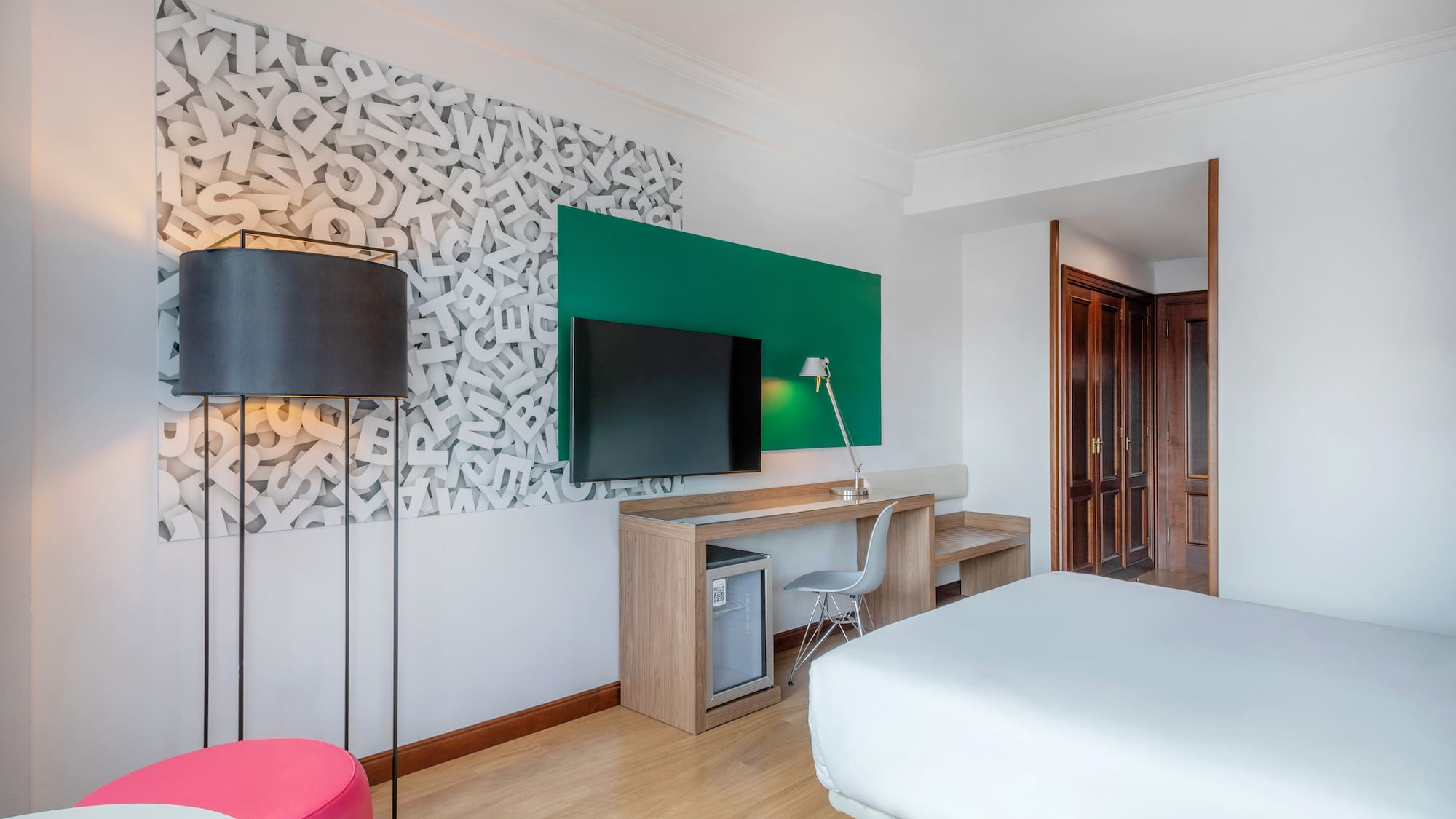 Modern hotel room with TV, desk, bed, and unique alphabet wall art.
