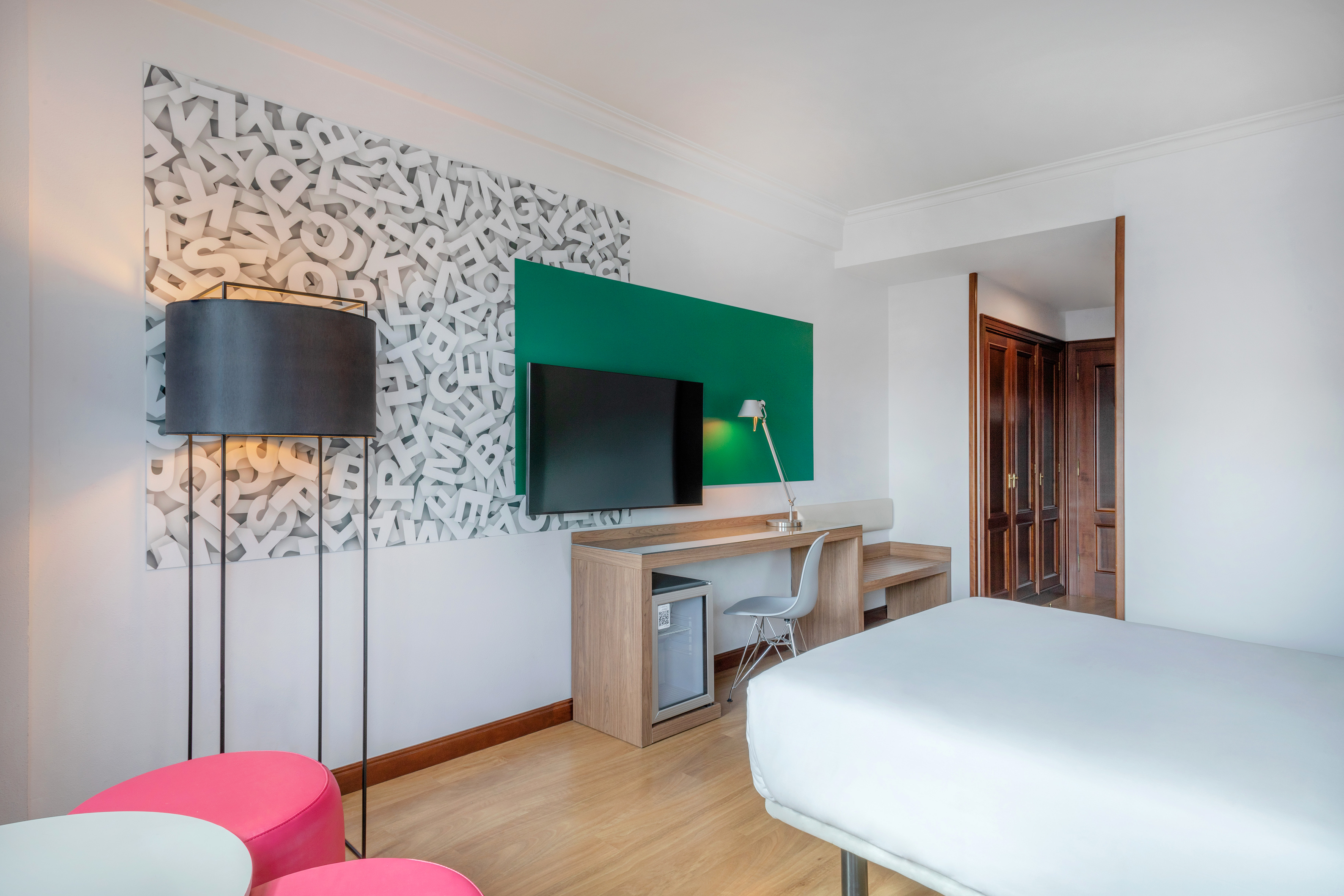 Modern hotel room with TV, desk, bed, and unique alphabet wall art.