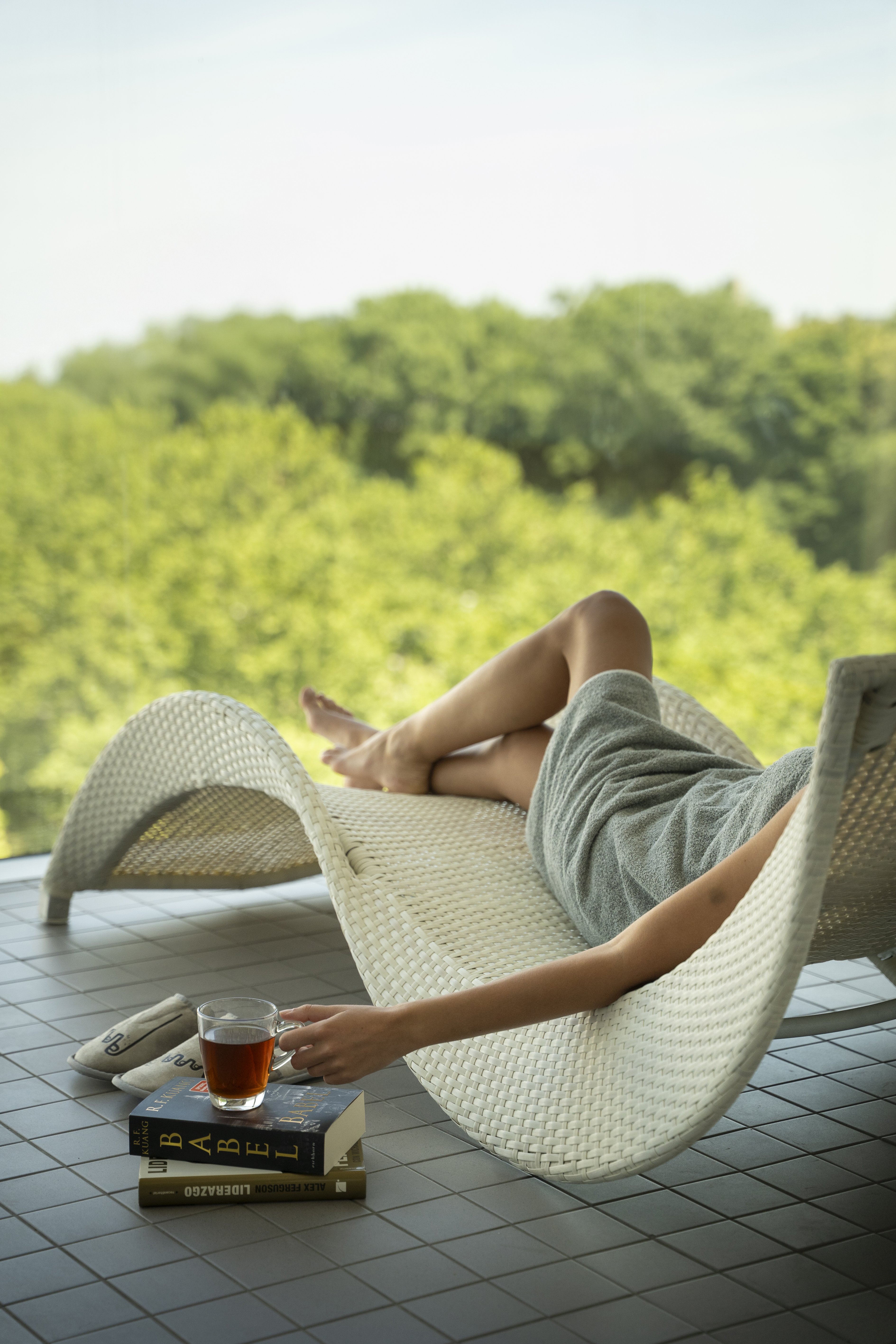 Person unwinding on spa lounge chair with tea, book, green forest view.