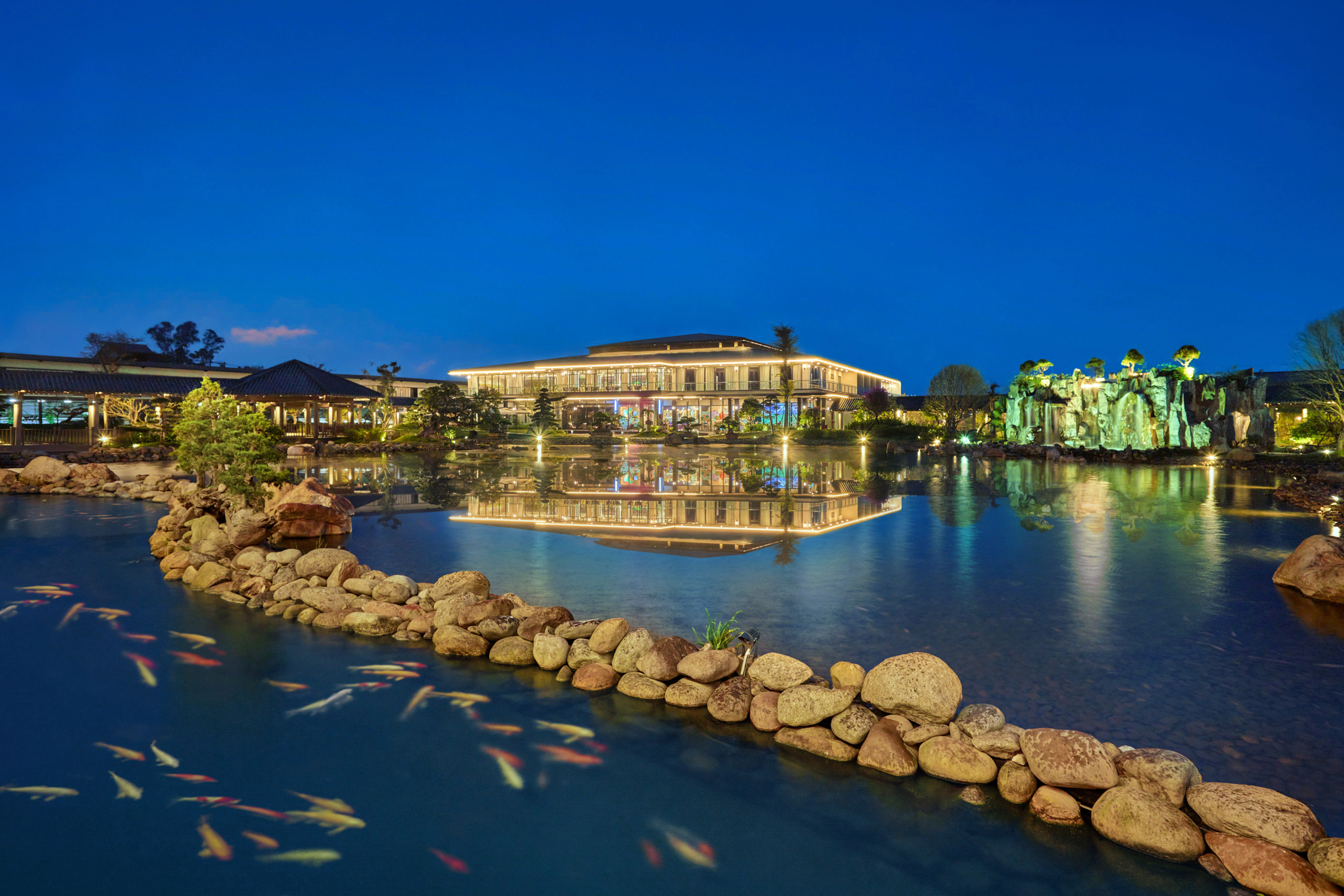 Illuminated resort reflecting in koi pond at night with a rocky waterfall.
